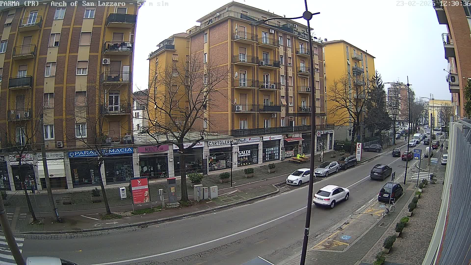 A street scene on an overcast day shows a multi-story yellow building with shops on the ground floor, parked cars along the curb, and several vehicles driving on the road.