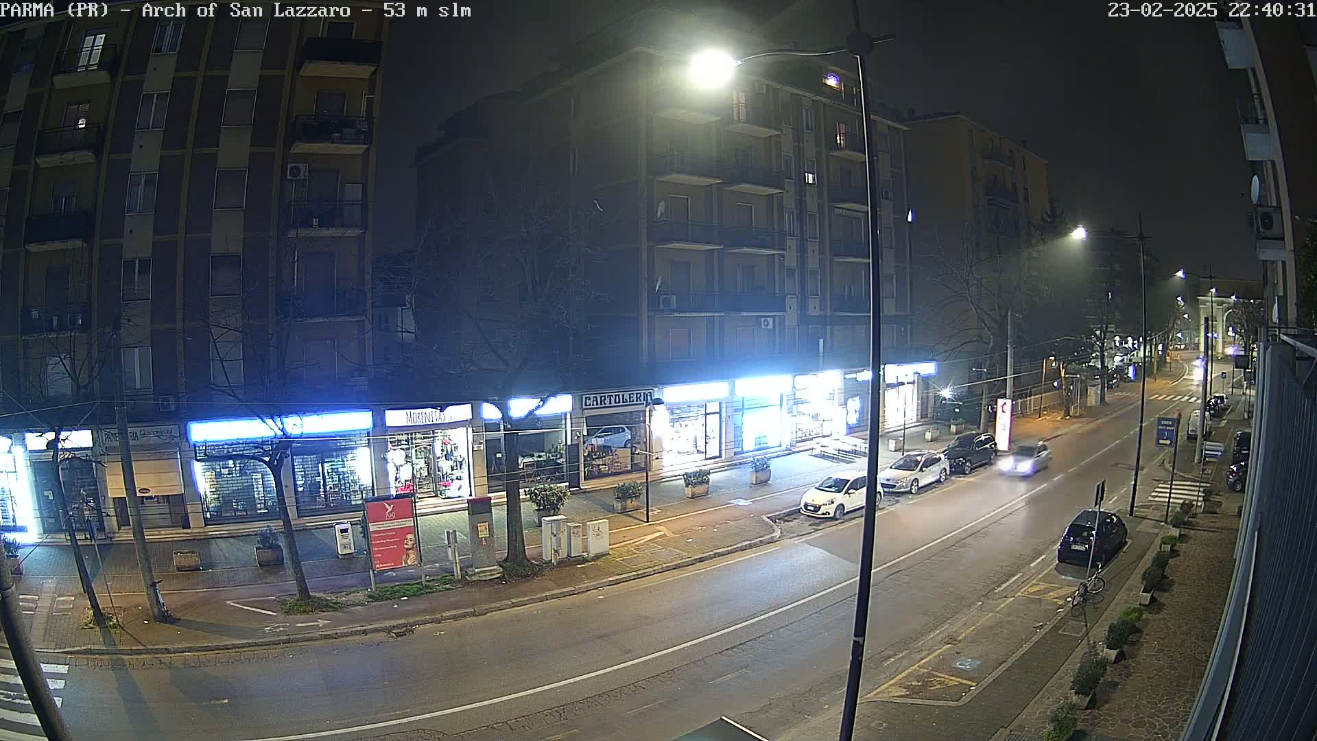 A nighttime street scene shows several parked cars and a few moving vehicles on a road lined with shops and a multi-story apartment building under streetlights.