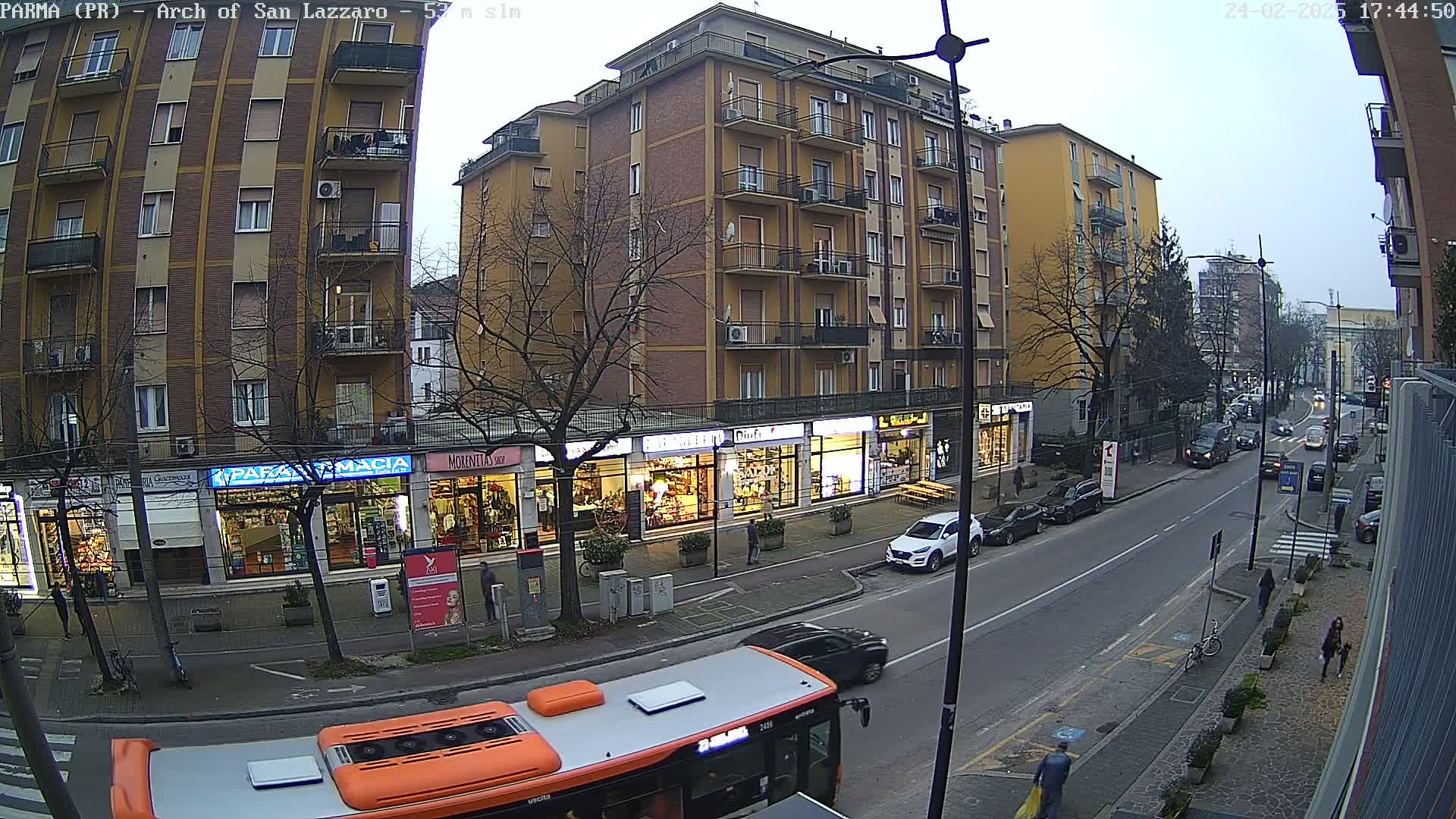 A street scene shows a multi-story building with shops on the ground floor, cars parked along the side of the road, and a bus in the foreground, under an overcast sky.