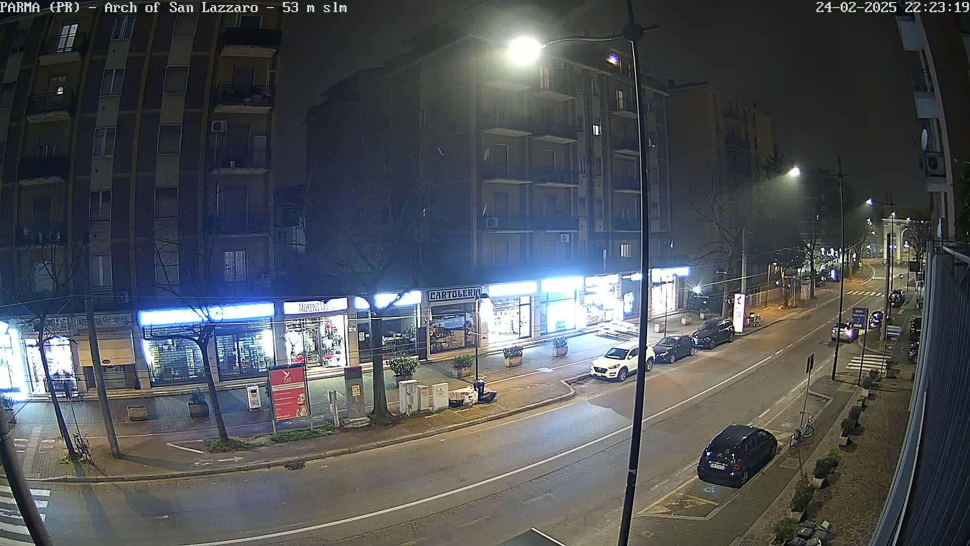 A nighttime street scene shows several parked cars, a few buildings with illuminated storefronts, and some streetlights illuminating the road.