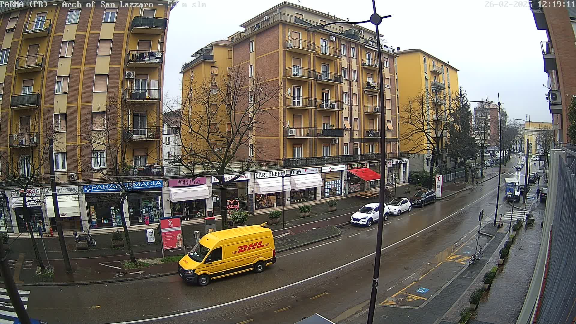 A wet city street scene shows a yellow DHL delivery van, several parked cars, and a multi-story brick building with shops on the ground floor under a gray sky.