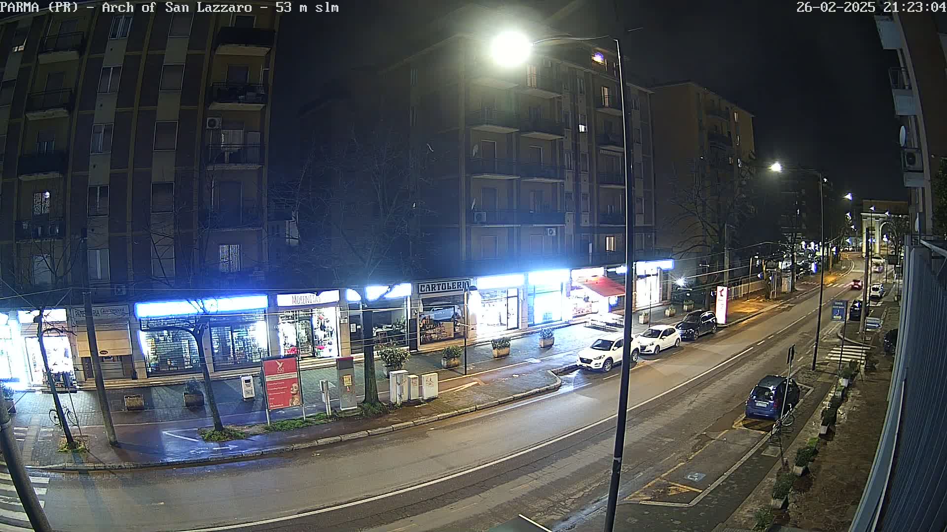 A nighttime street scene shows a row of shops and parked cars along a road under streetlights, with a few cars driving by.