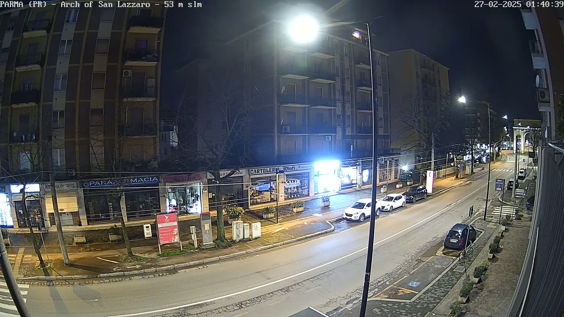 A nighttime street scene shows a mostly empty road lined with shops and apartment buildings under bright streetlights.