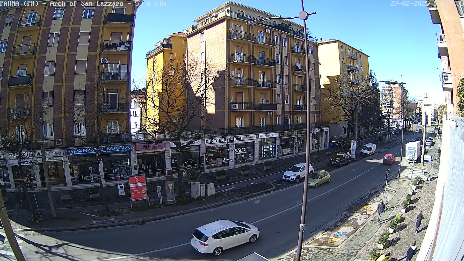 A sunny city street scene shows several multi-story buildings, shops, and cars, with pedestrians walking on a sidewalk.