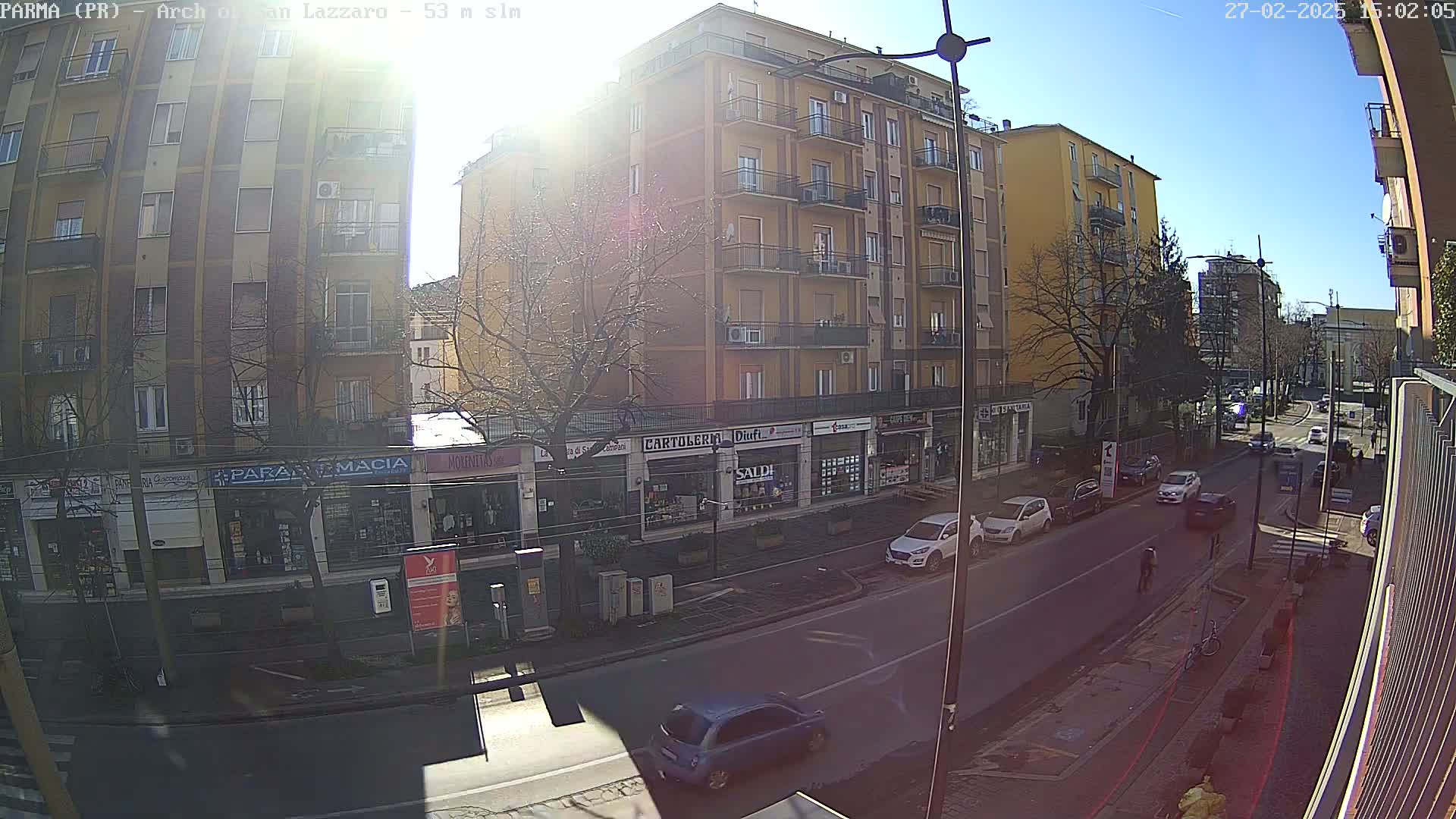 A sunny street scene shows a multi-story building with shops on the ground floor, several parked cars, and a few pedestrians, all under a clear blue sky.