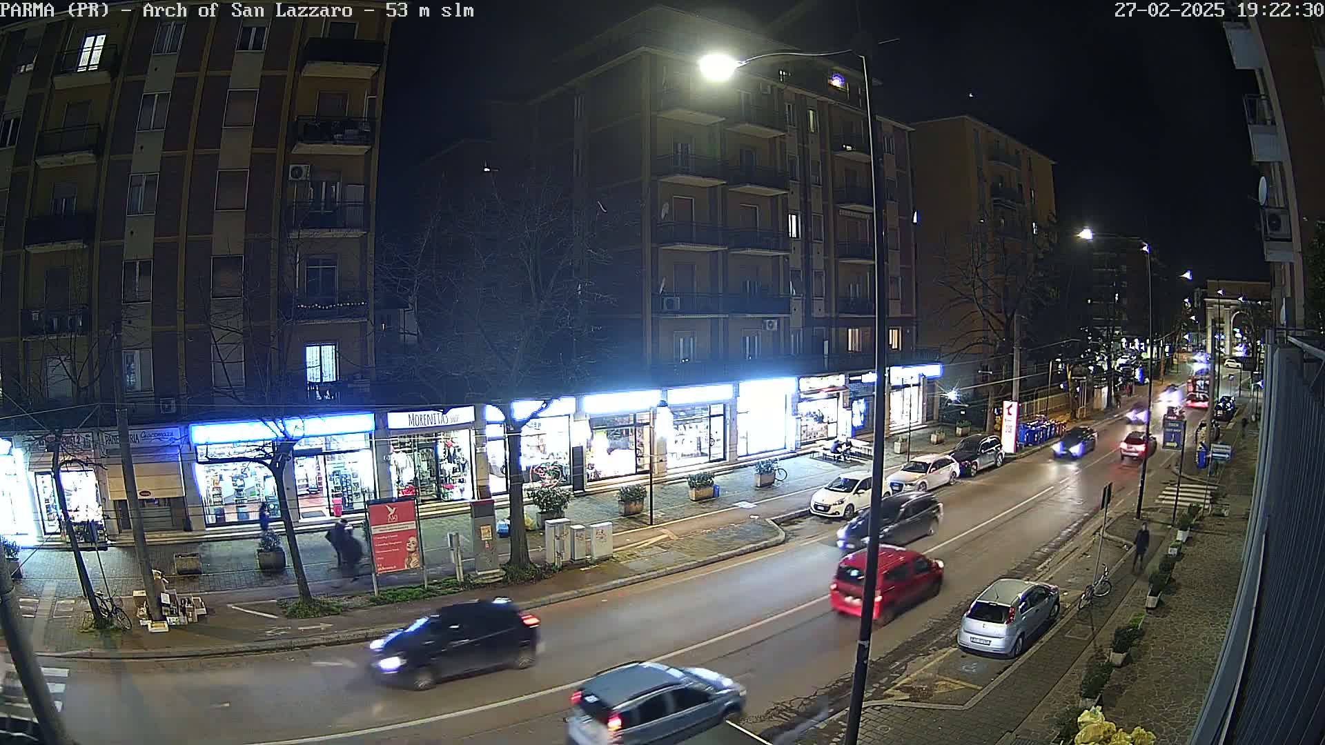 A nighttime street scene shows several cars driving on a city street lined with shops and apartment buildings under a clear, dark sky.