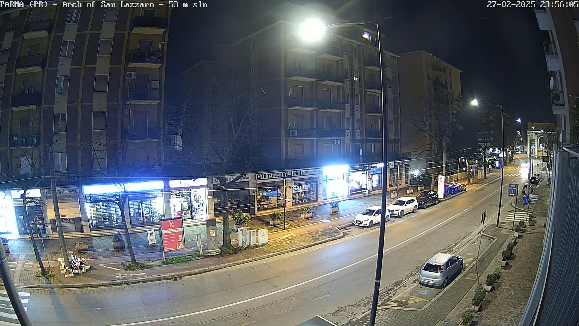 A nighttime street scene shows several parked cars along a roadway lined with shops and apartment buildings under streetlights.