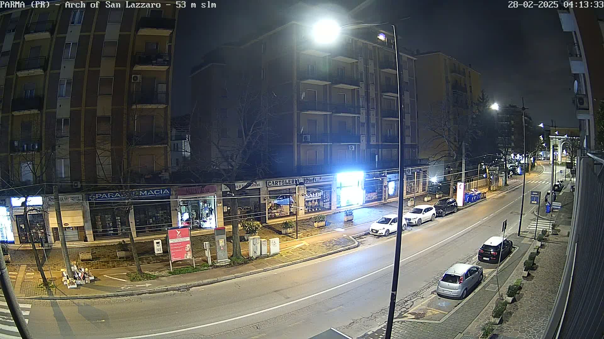A nighttime street scene shows parked cars along a multi-story building with shops on the ground floor under streetlights.