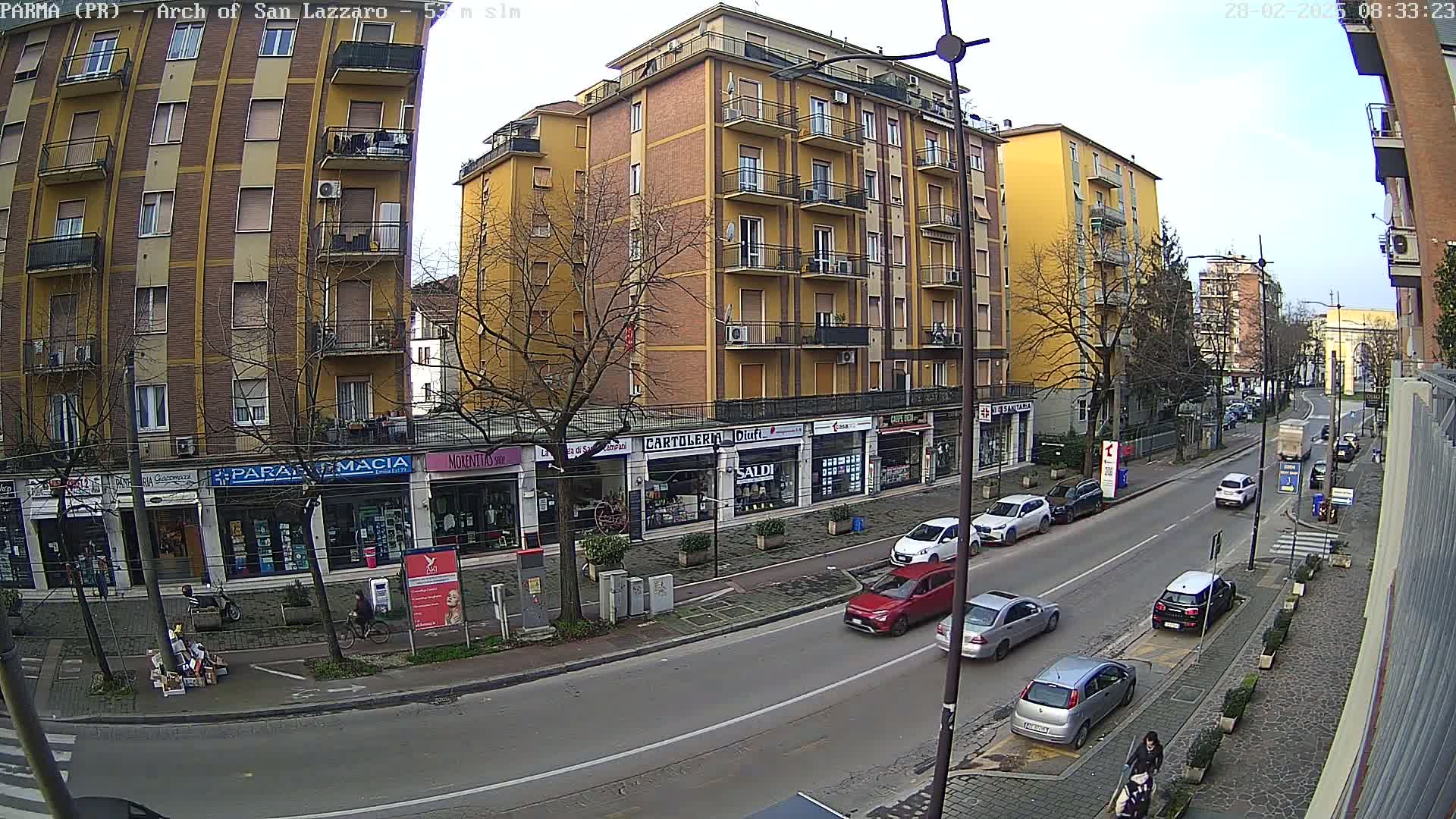 A mostly sunny street scene shows a multi-story yellow building with shops on the ground floor, and several cars parked and driving along a road lined with bare trees.