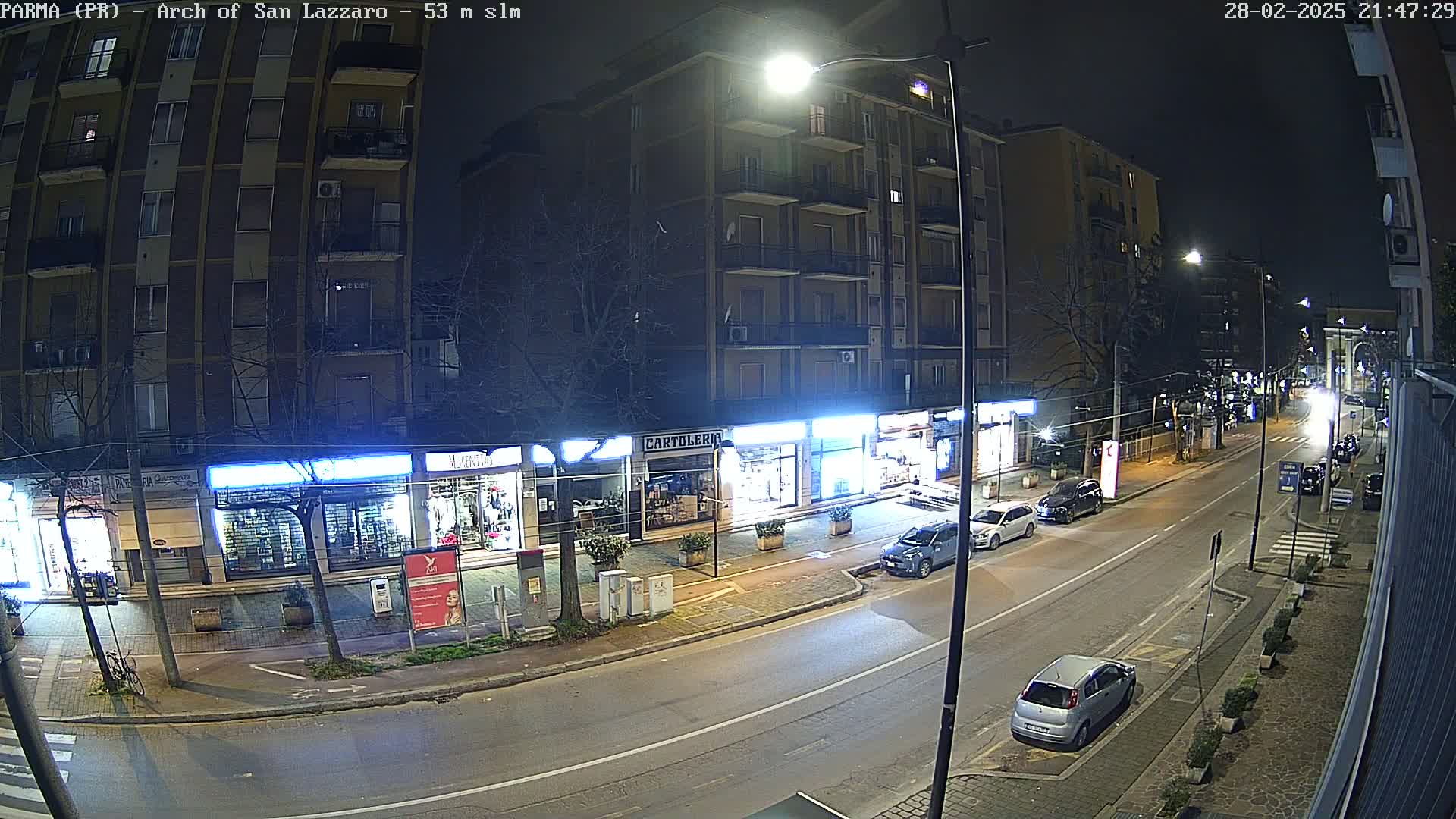 A nighttime street scene shows a row of shops and buildings with cars parked along the curb and a few cars driving on the road under streetlights.