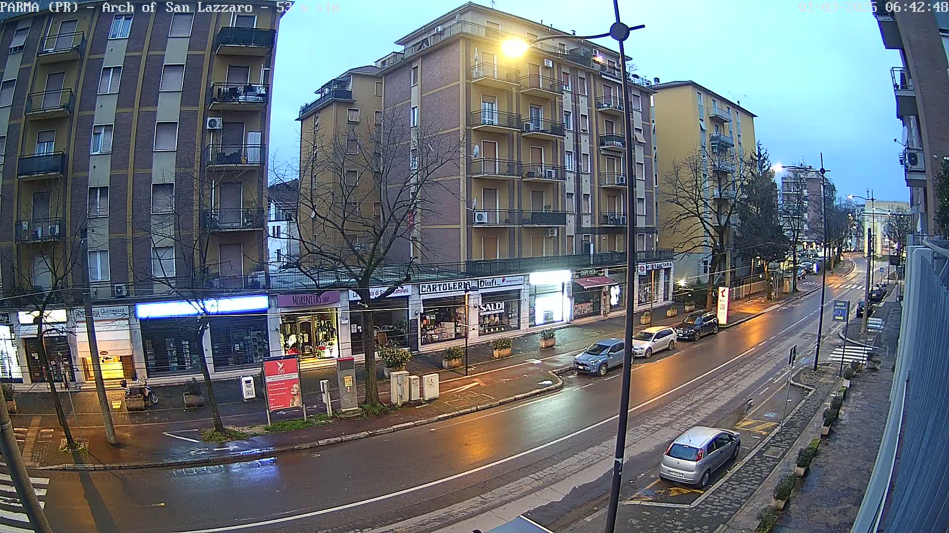 A wet city street at dusk, lined with multi-story buildings and shops, shows several parked cars and a few driving down the street.