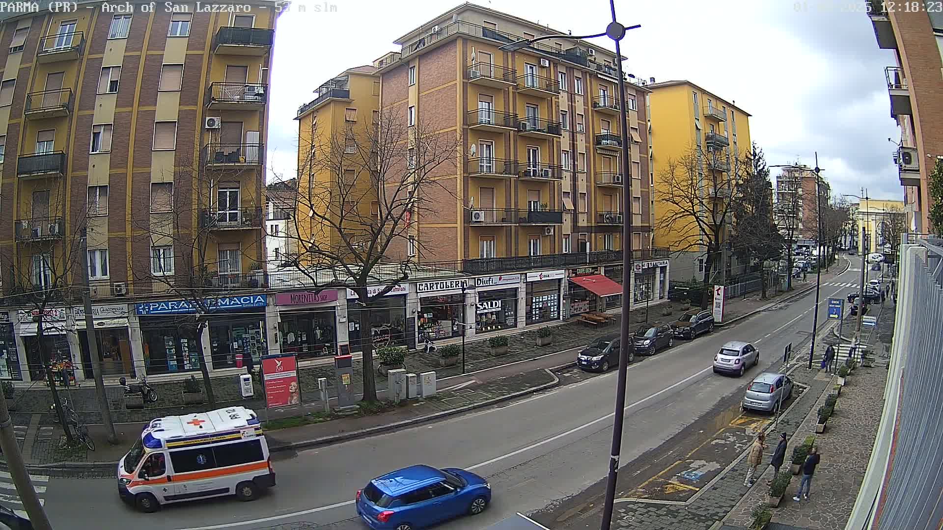 A street scene shows a multi-story yellow building with shops on the ground floor, several cars parked and driving along the street, and an ambulance, all under an overcast sky.