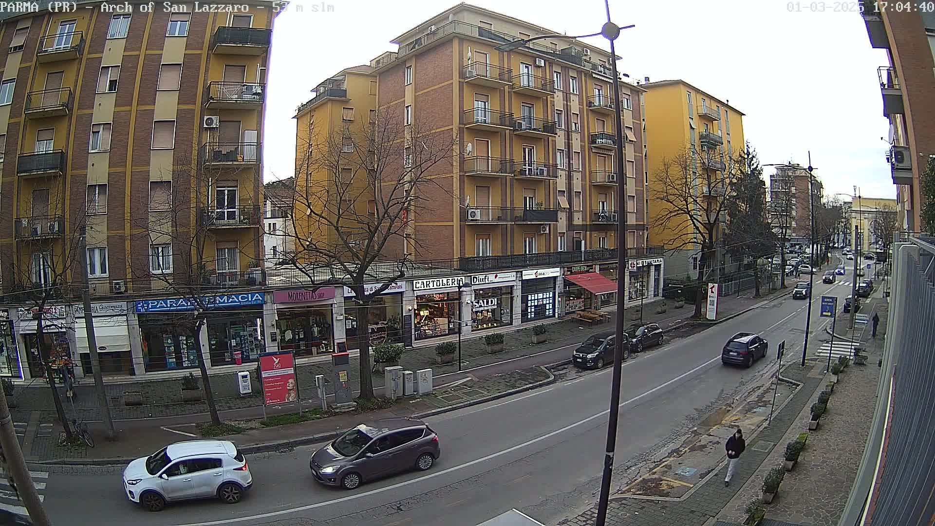A street scene on an overcast day shows a multi-story building with shops on the ground floor, several cars parked and driving along the road, and a pedestrian walking on the sidewalk.