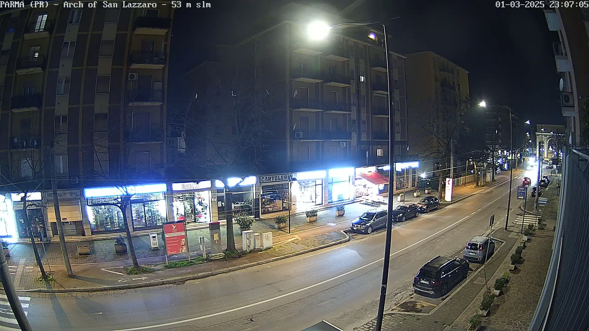 A nighttime street scene shows several parked cars and a few shops along a roadway under bright streetlights.
