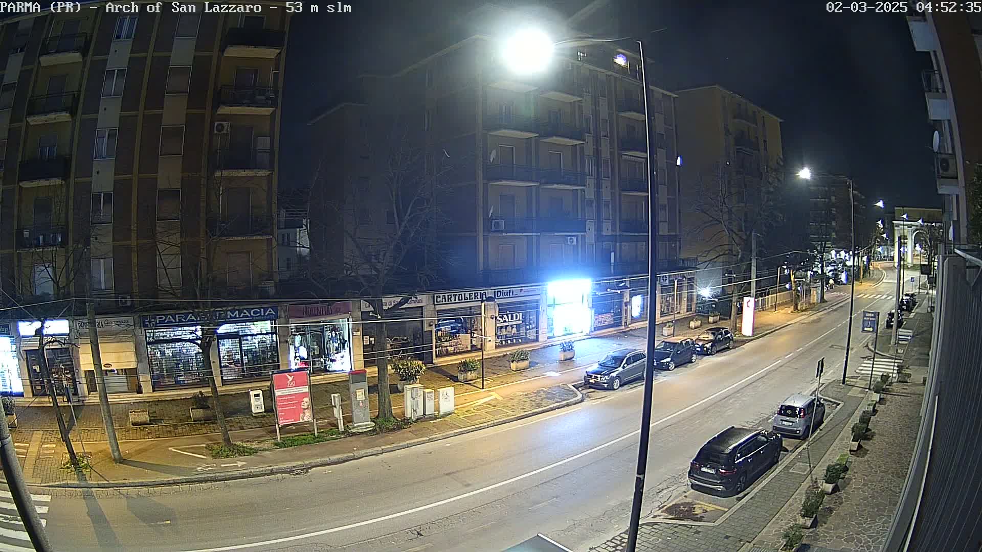 A nighttime street scene shows several parked cars along a city road lined with shops and buildings under streetlights.