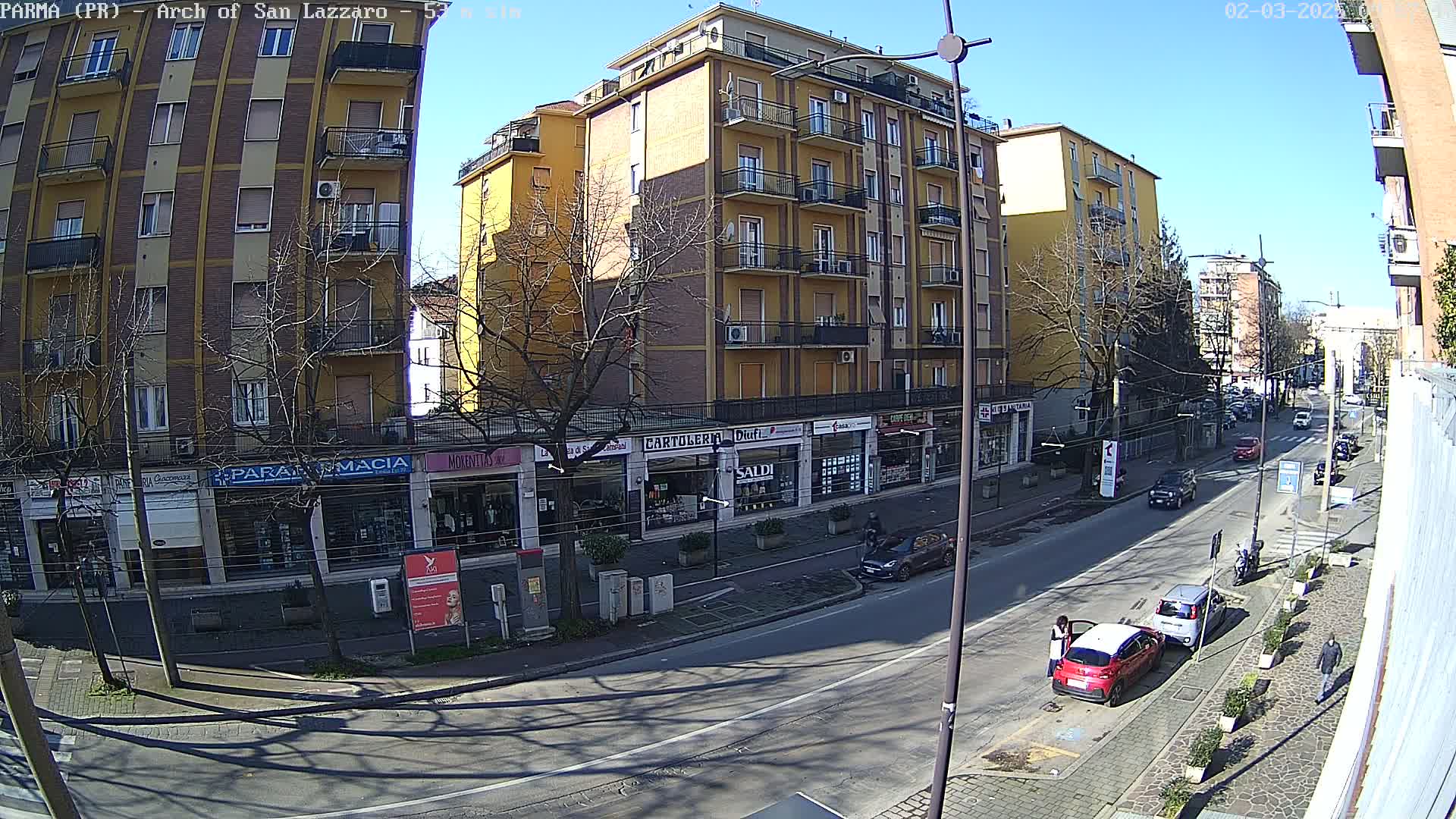 A sunny day on a city street, showing several multi-story buildings with shops on the ground floor, a few cars parked along the curb, and sparse pedestrian activity.
