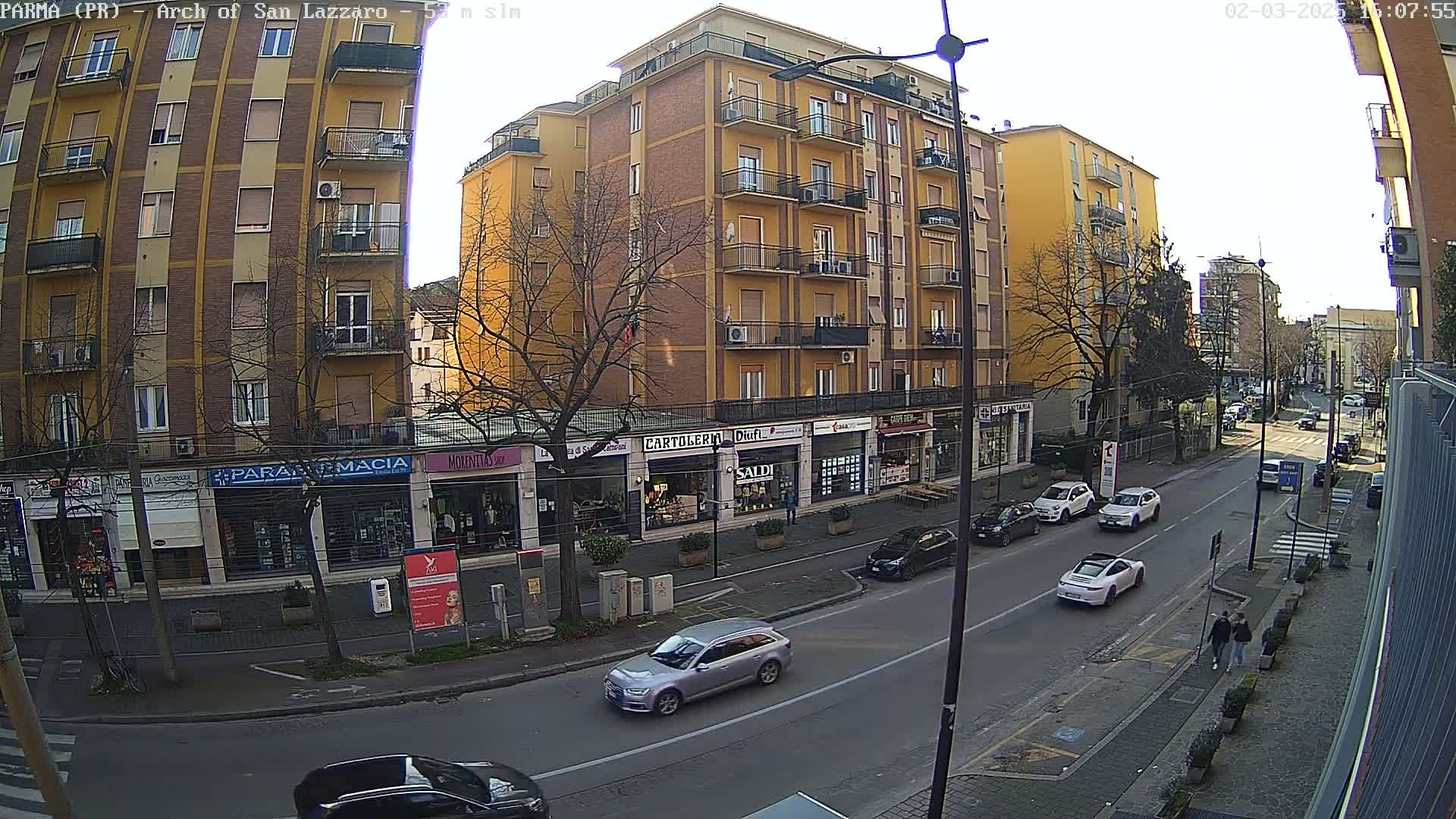 A sunny city street scene shows several cars parked and driving past multi-story buildings with ground-level shops.