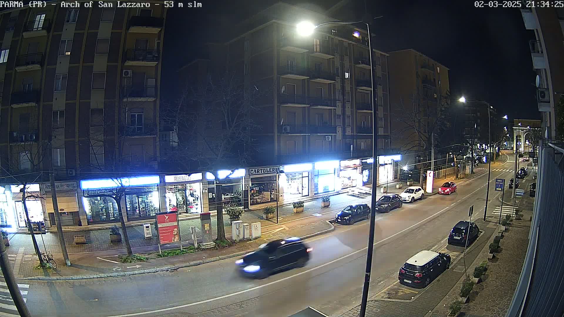 A nighttime street scene shows several cars parked and driving along a road lined with storefronts and a multi-story building.