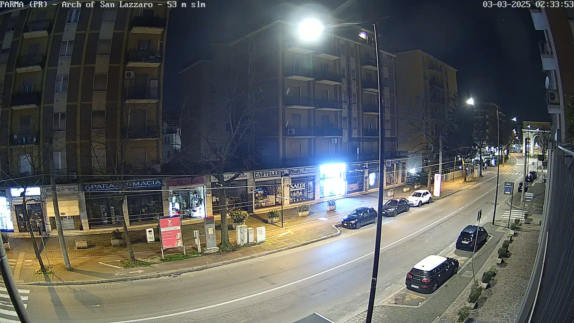 A nighttime street scene shows a relatively empty road with several cars parked along the sides, in front of multi-story buildings with ground-floor shops.