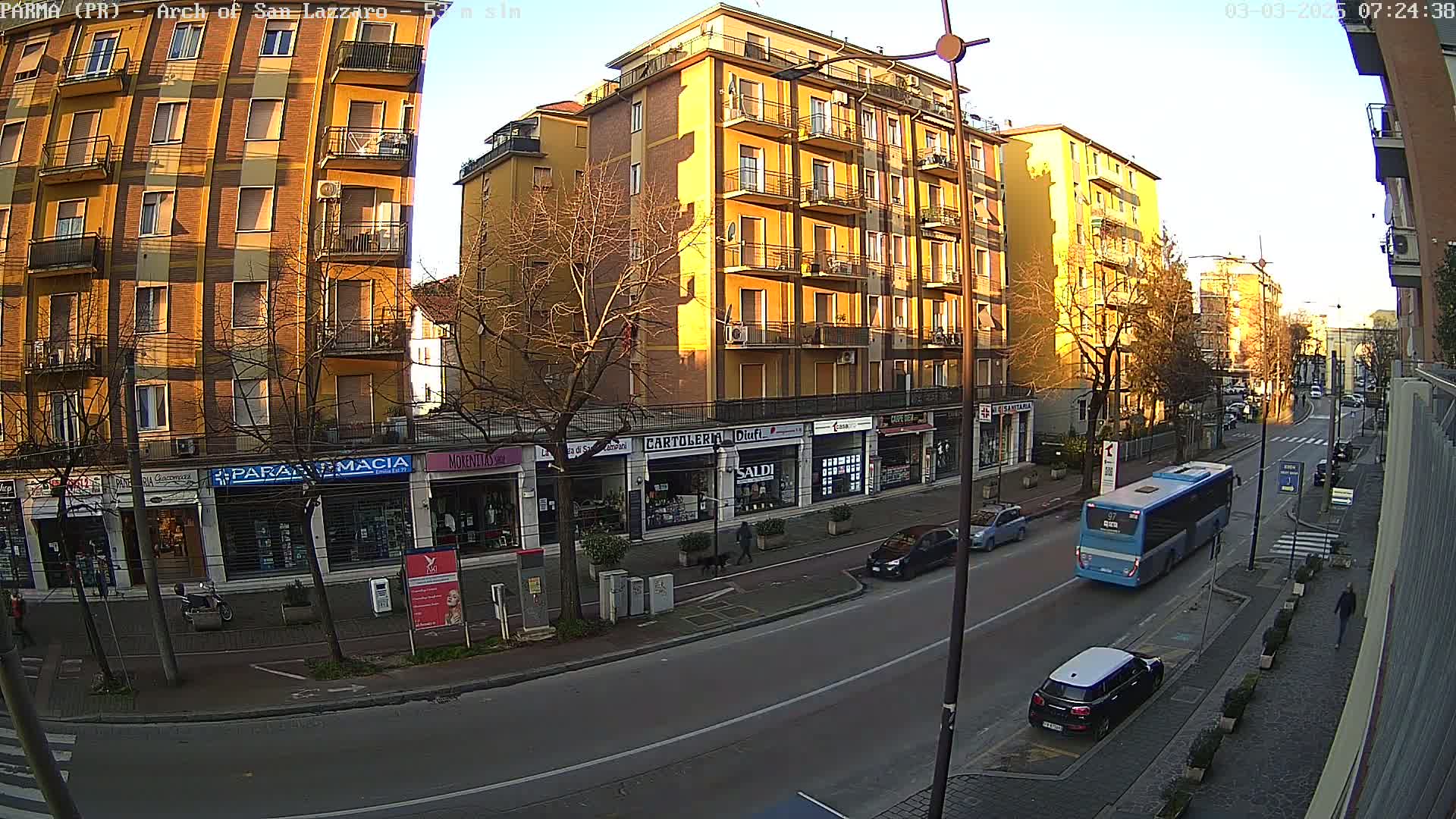 A sunny street scene shows a multi-story yellow building with shops on the ground floor, several cars and a bus on the road, and bare trees lining the sidewalks.