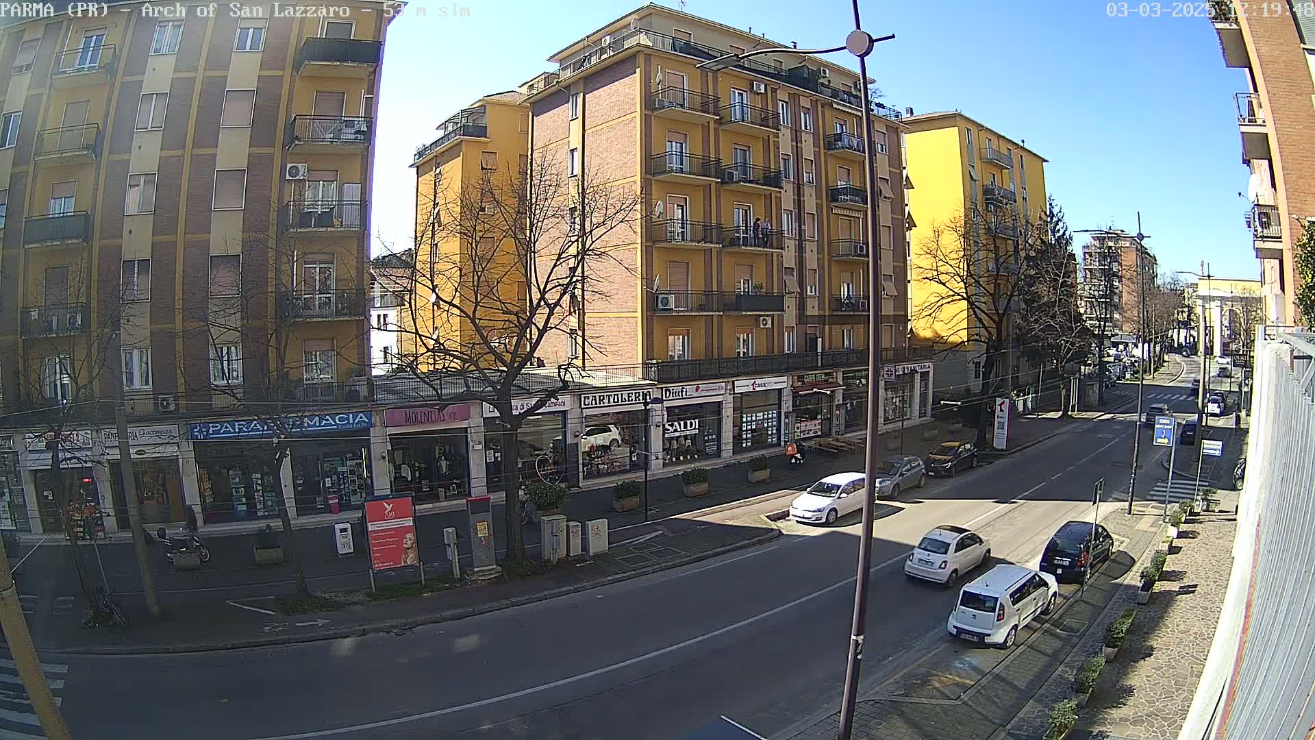 A sunny street scene shows a multi-story yellow building with shops on the ground floor, several cars parked and driving on the street, and bare trees lining the road.