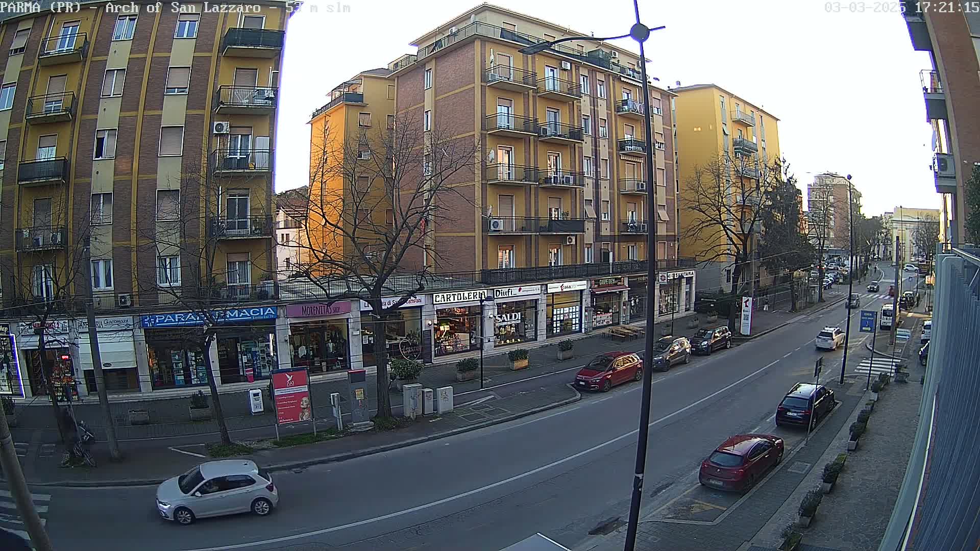 A street scene shows a row of multi-story buildings with shops on the ground floor under a partly sunny sky.