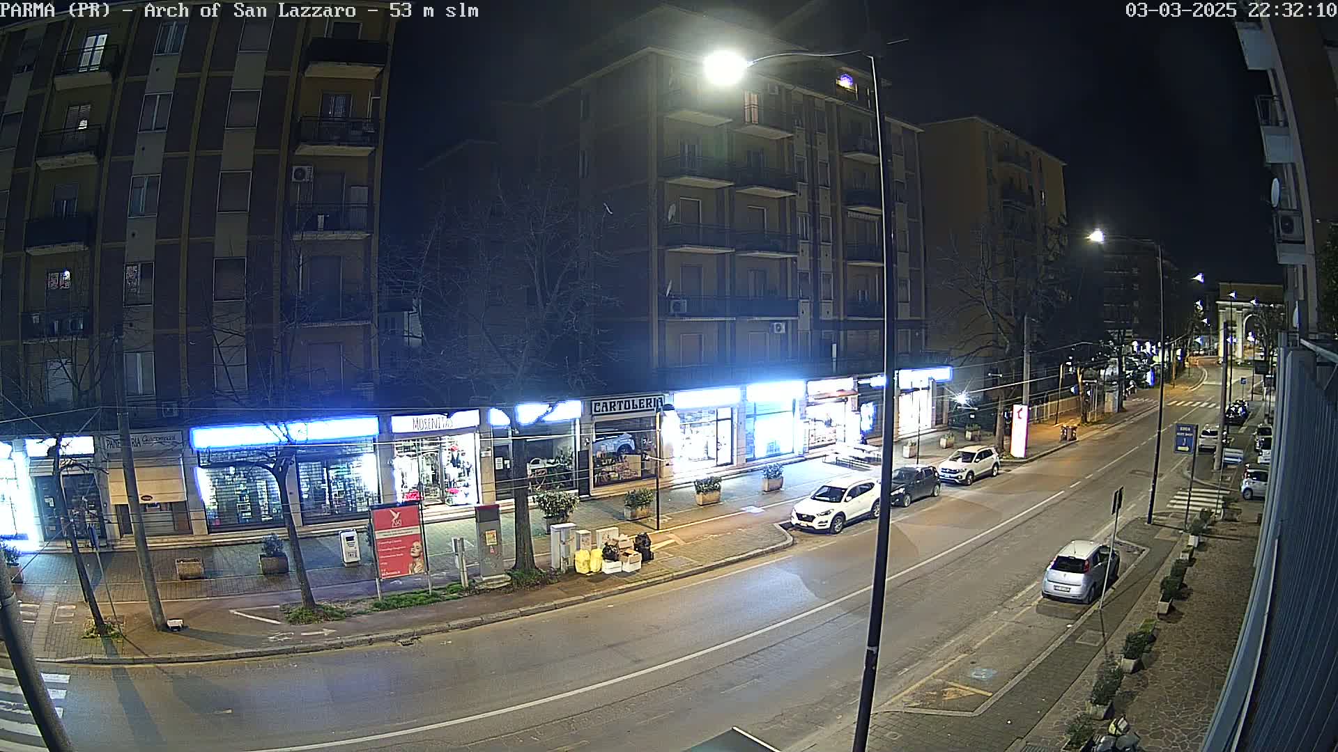 A nighttime street scene shows several parked cars alongside a multi-story building with illuminated storefronts.
