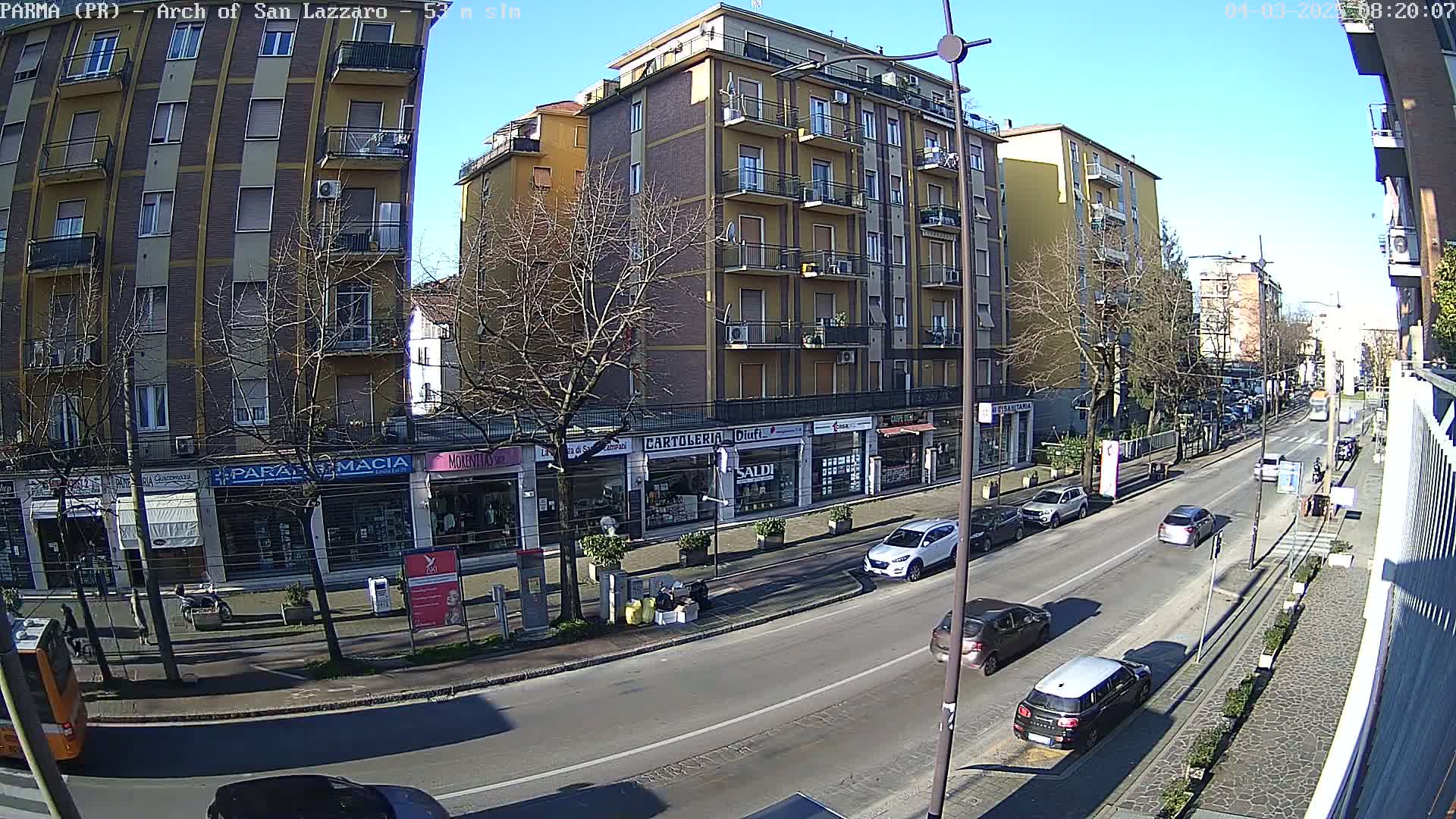 A sunny day reveals a city street scene with several cars parked and driving along a roadway lined with shops and a multi-story apartment building.