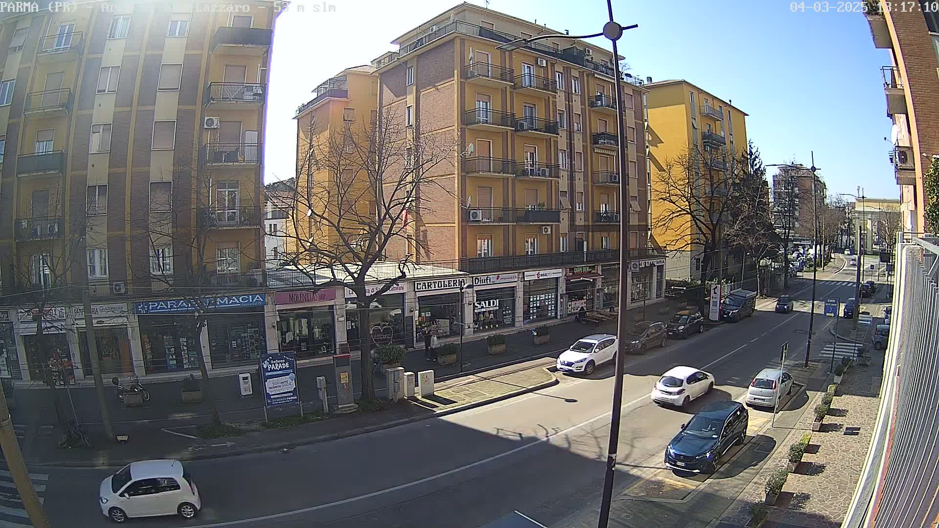 A sunny street scene shows several cars parked and driving along a two-lane road in front of multi-story buildings with shops on the ground floor.