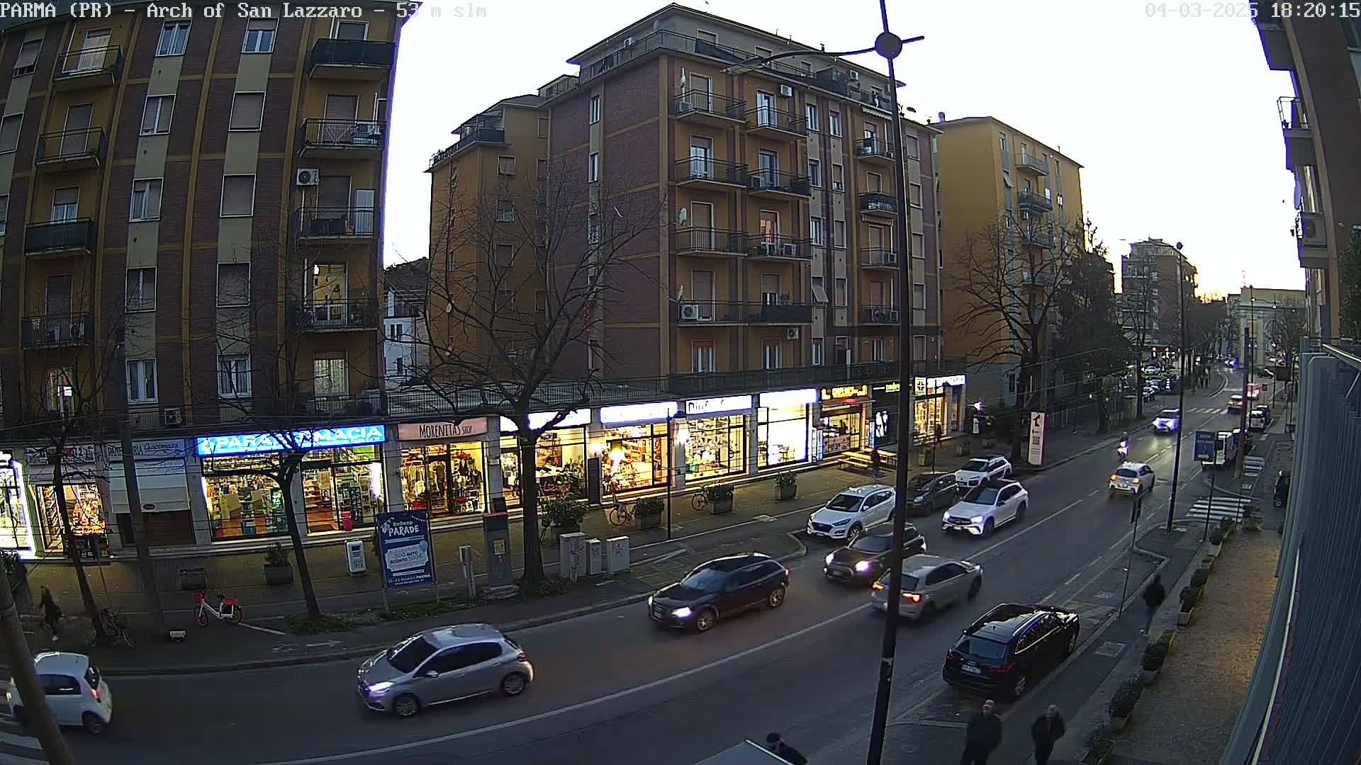 A street scene at dusk shows several cars driving on a road lined with multi-story buildings and storefronts.