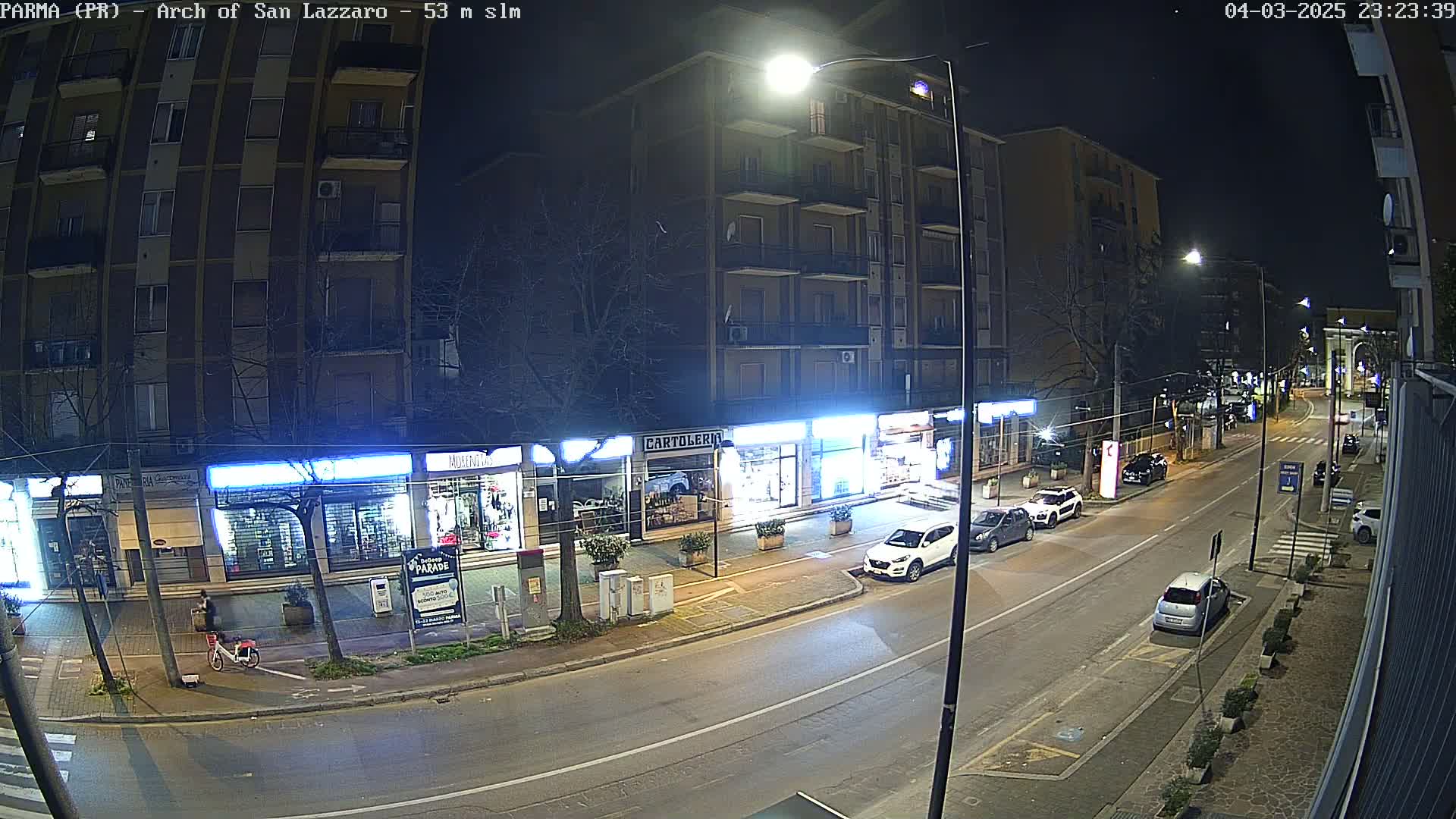 A nighttime street scene shows a row of shops and buildings under streetlights with several cars parked along the curb and a few driving down the road.