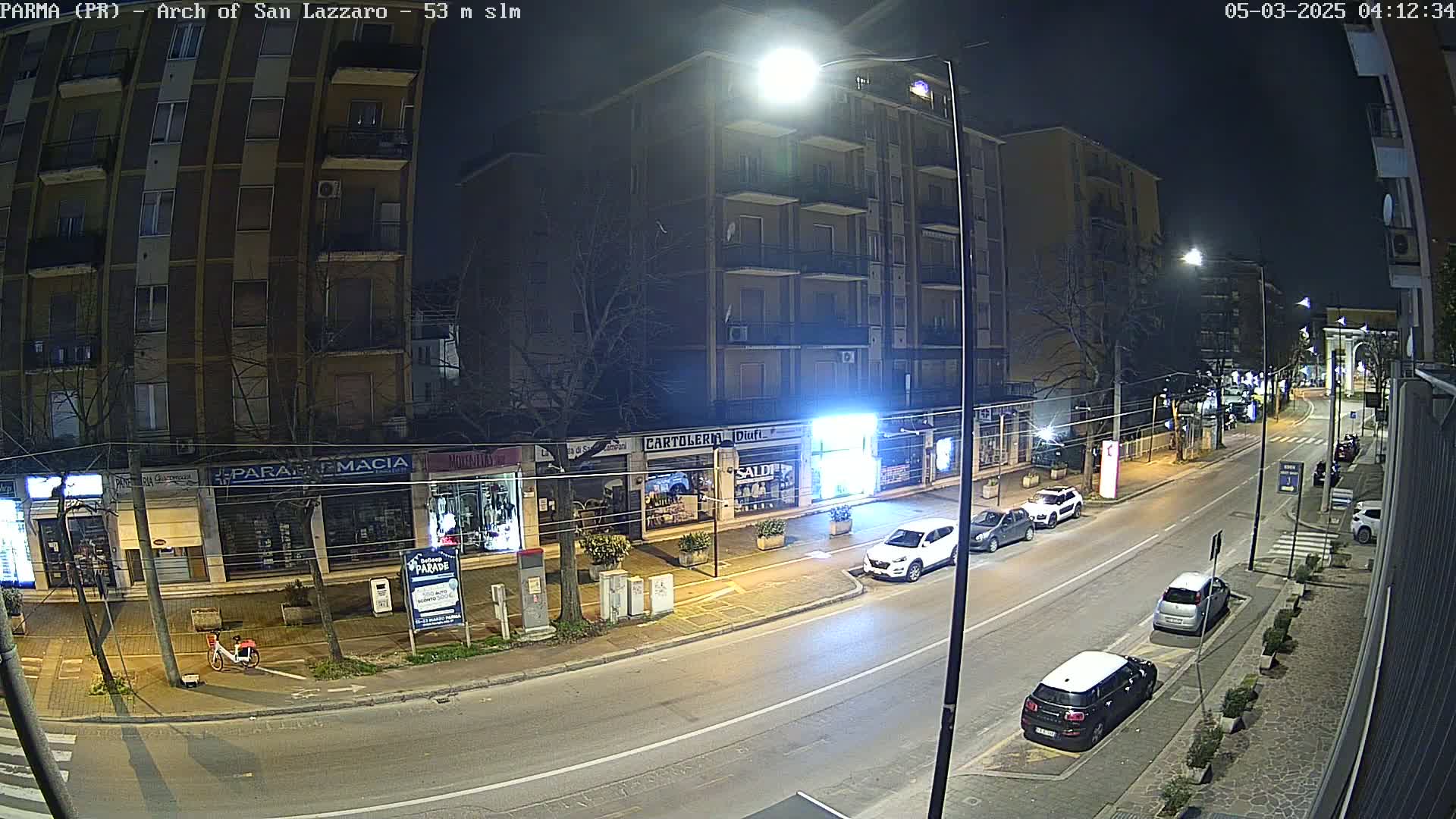 A nighttime street scene shows a multi-story building with shops on the ground floor, several cars parked along the curb, and a few more driving down the street.