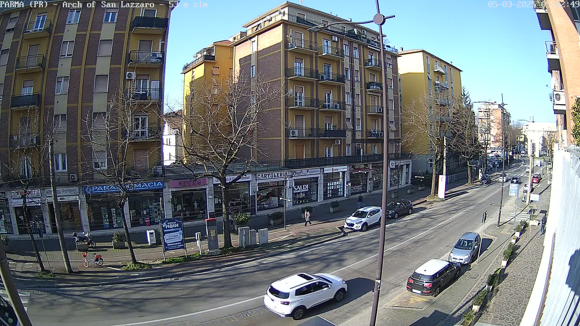 A sunny street scene shows several cars parked and driving along a multi-lane road lined with multi-story buildings that have ground-floor shops.