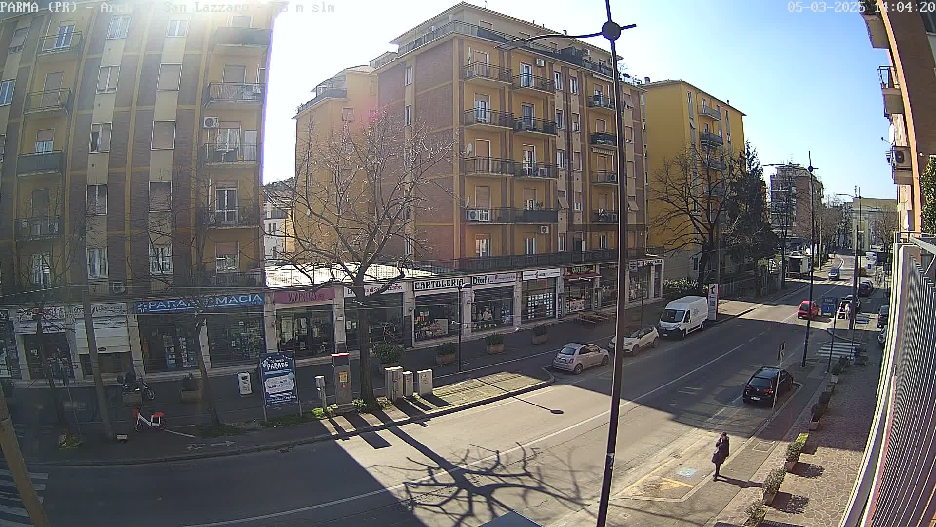 A sunny street scene shows a row of buildings with shops on the ground floor, several cars parked along the street, and a few pedestrians walking.