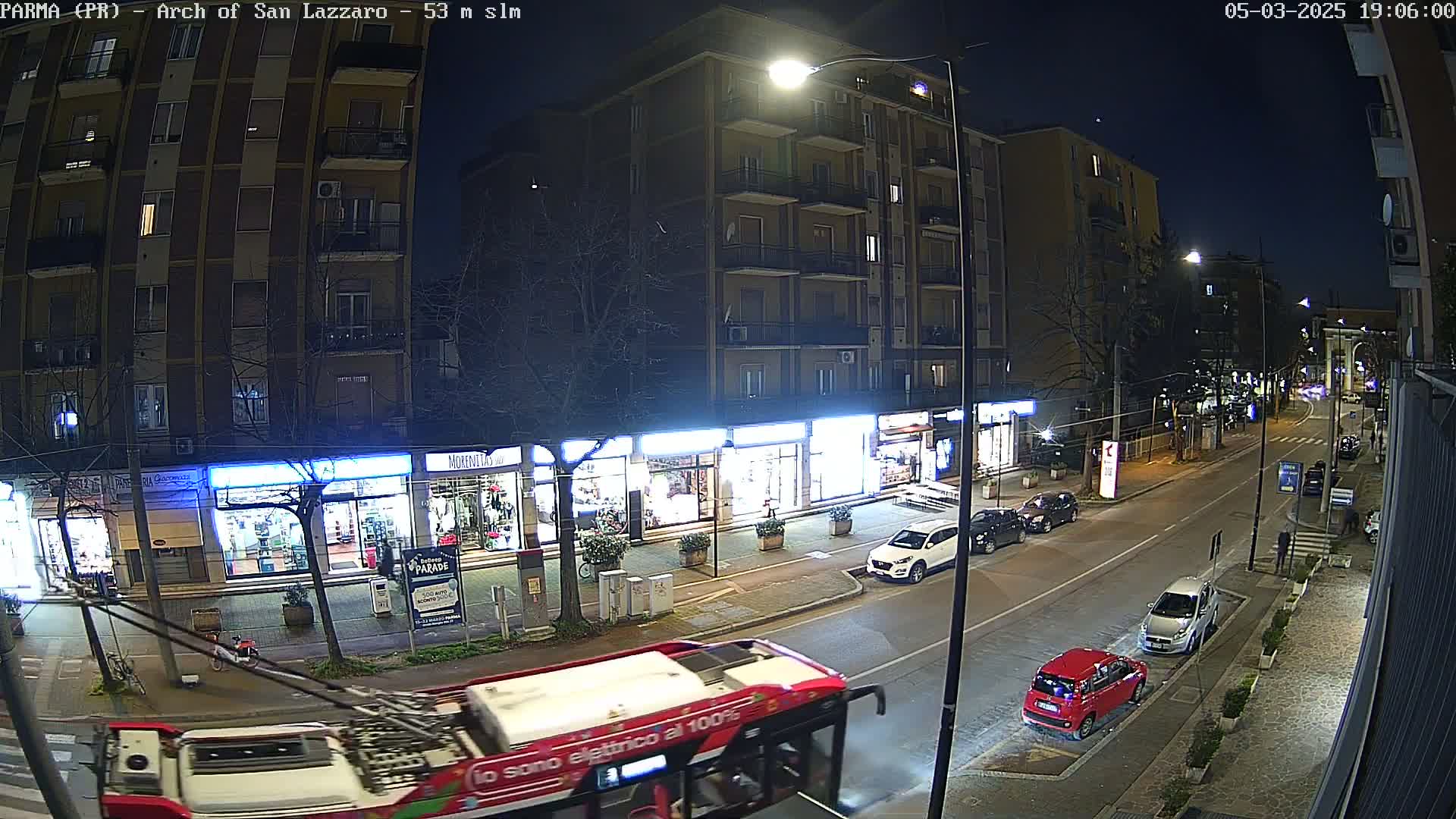 A nighttime street scene shows a red double-decker bus approaching a row of shops and parked cars under streetlights.