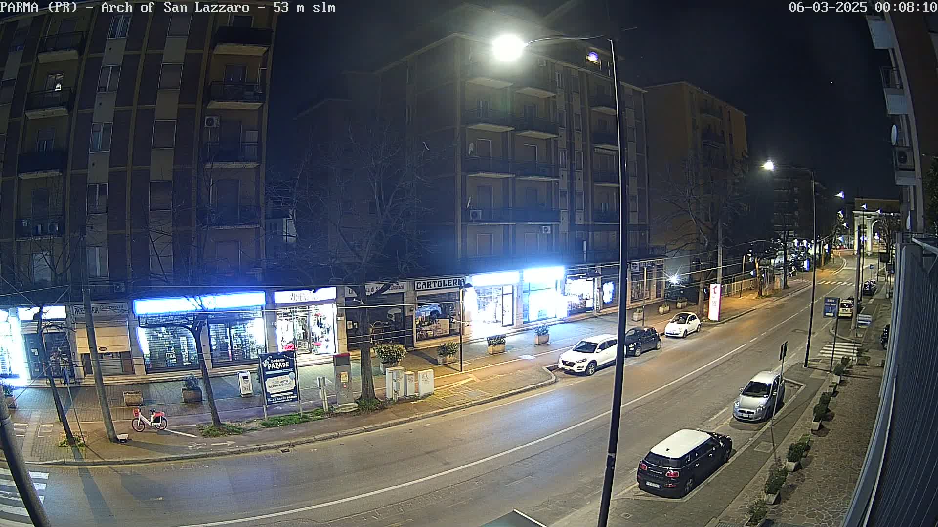 A nighttime street scene shows several cars parked along the sides of a multi-story building with shops on the ground floor, under streetlights.