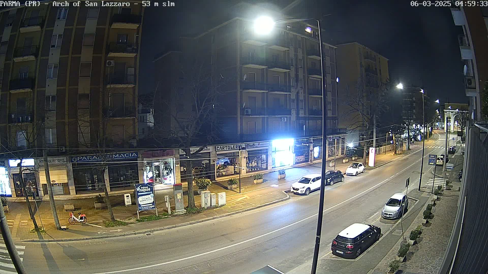 A nighttime street scene shows a mostly empty road lined with shops and apartment buildings, with several cars parked along the curb and a few driving down the road.