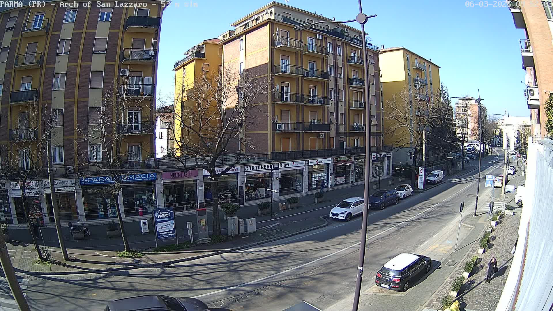 A sunny city street scene shows several multi-story buildings with shops on the ground floor, parked cars along the side, and a few pedestrians walking on the sidewalk.