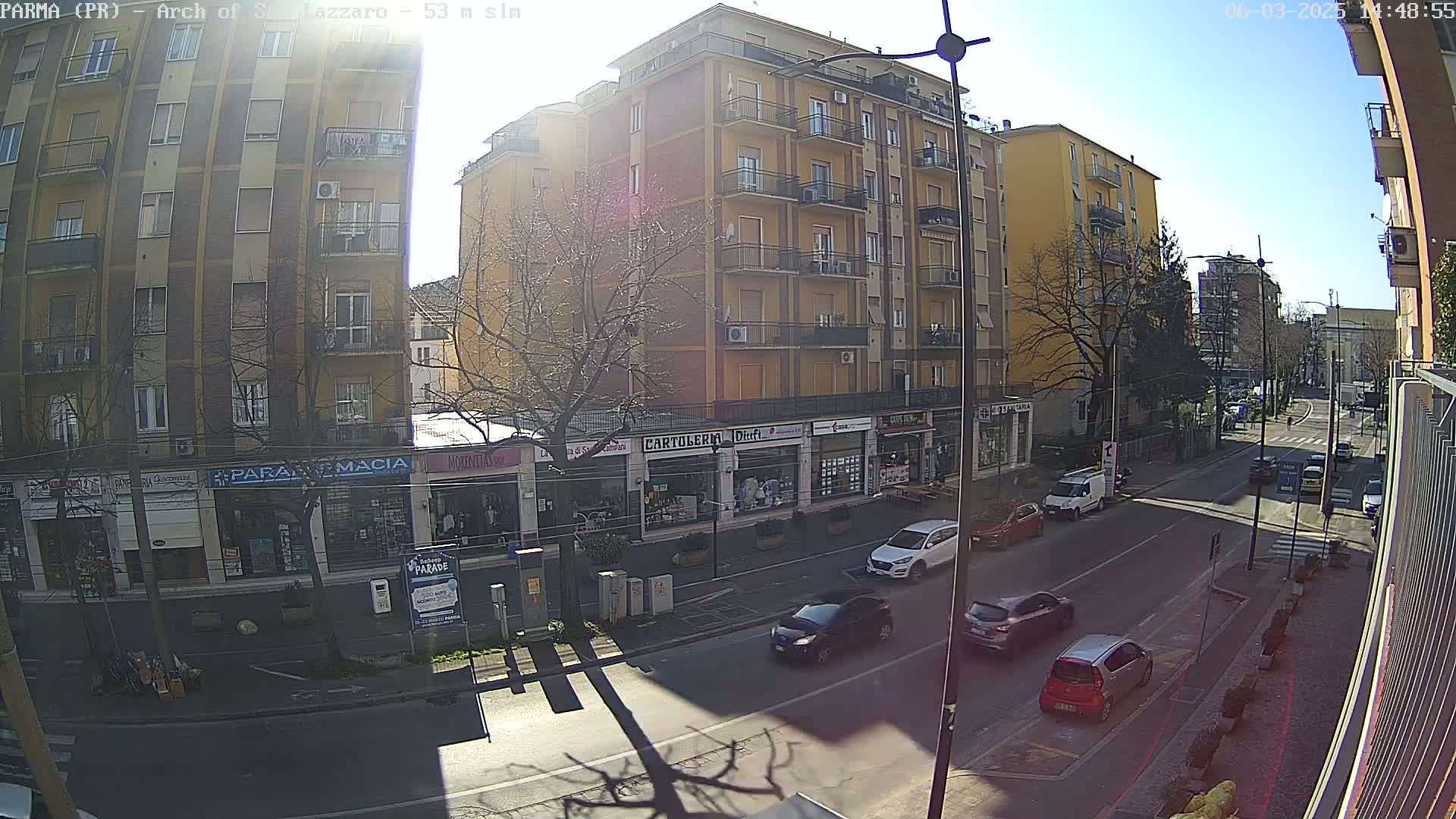 A sunny street scene shows a multi-story yellow building with shops on the ground floor, several cars parked and driving along the road, and bare trees.