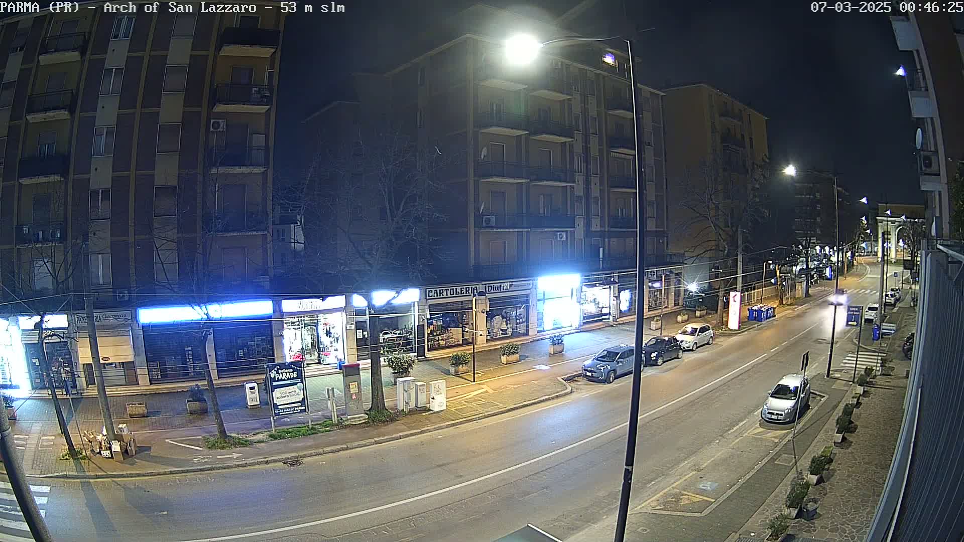 A nighttime street scene shows a mostly empty road with parked cars in front of shops and apartment buildings under streetlights.
