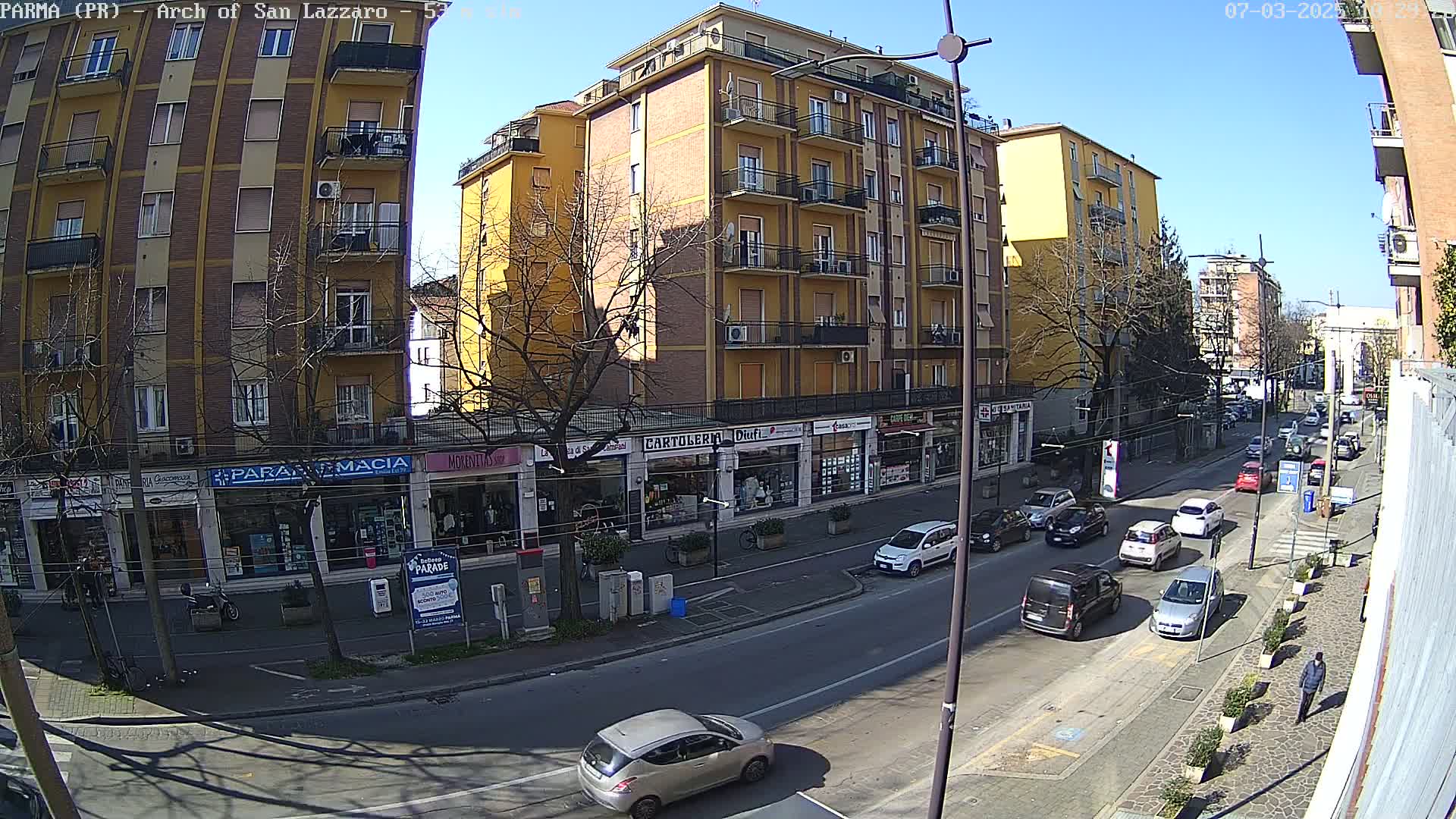 A sunny day on a city street lined with shops and apartment buildings, showing several cars driving down the road.