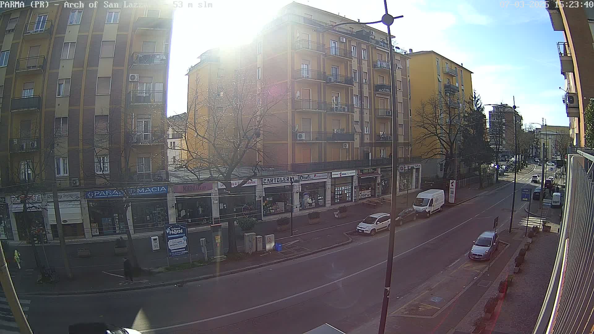 A sunny street scene shows a multi-story building with shops on the ground floor, several cars parked along the road, and pedestrians walking on the sidewalk.