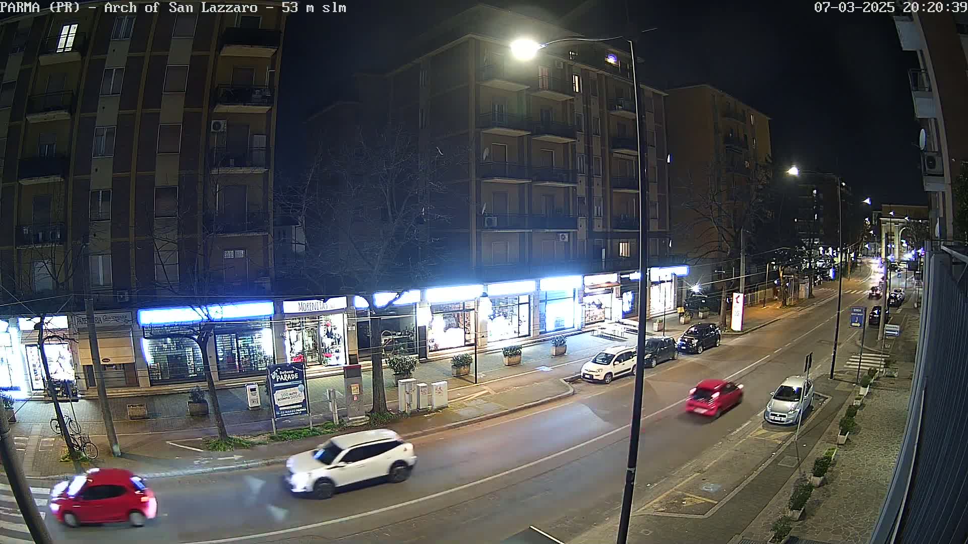 A nighttime street scene shows a multi-story building with storefronts on the ground floor, several cars parked and driving along the street, and a few pedestrians visible in the distance under clear skies.