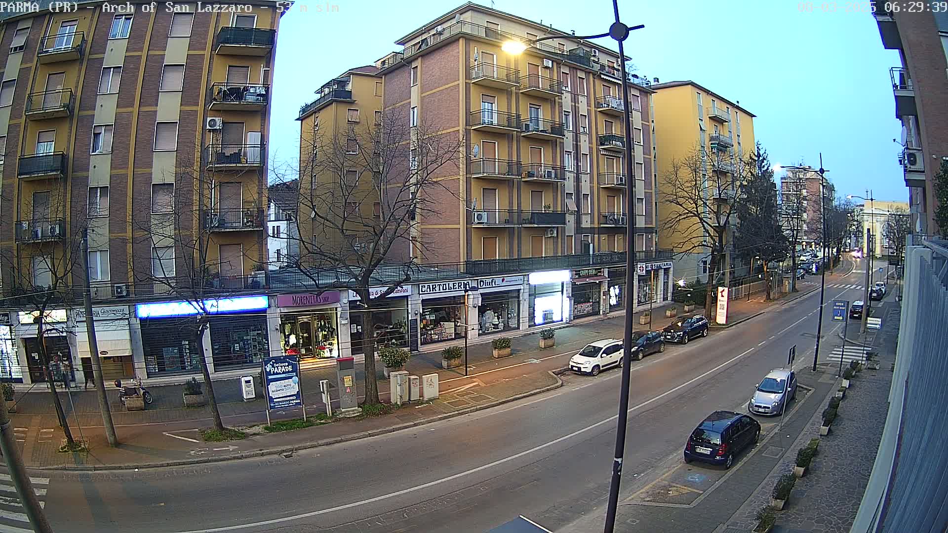 A street scene at twilight shows several parked cars along a two-lane road lined with multi-story buildings and shops.