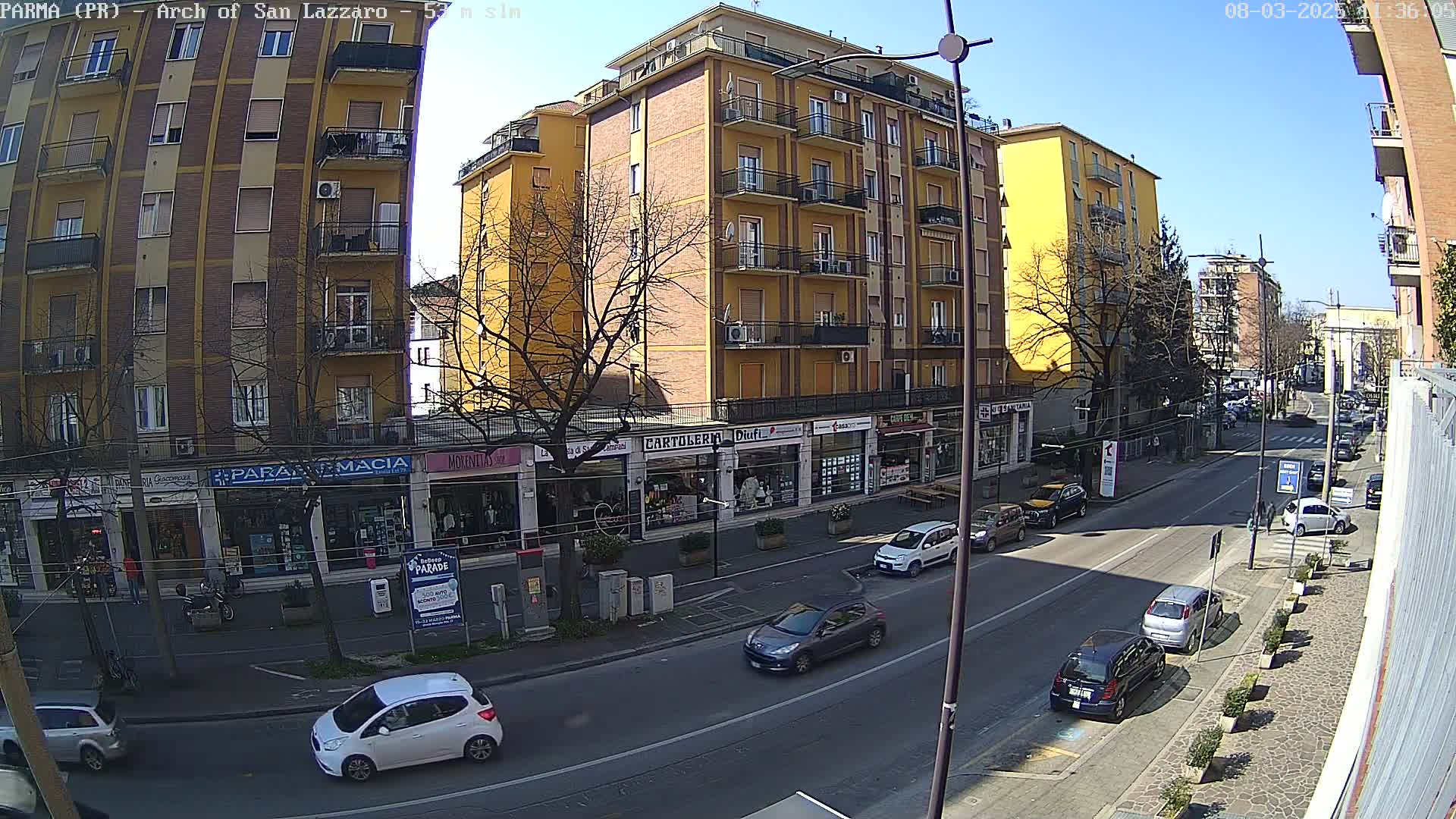 A sunny street scene shows several multi-story buildings, parked cars, and a few cars driving down a street lined with shops and trees.