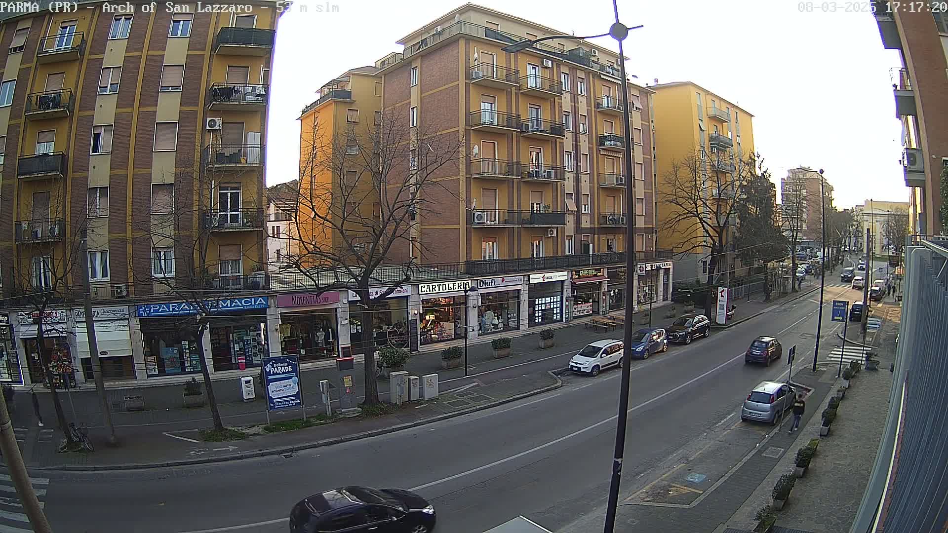 A street scene on a partly sunny day shows a row of multi-story buildings with shops on the ground floor, and several cars parked along the curb of a street with light traffic.