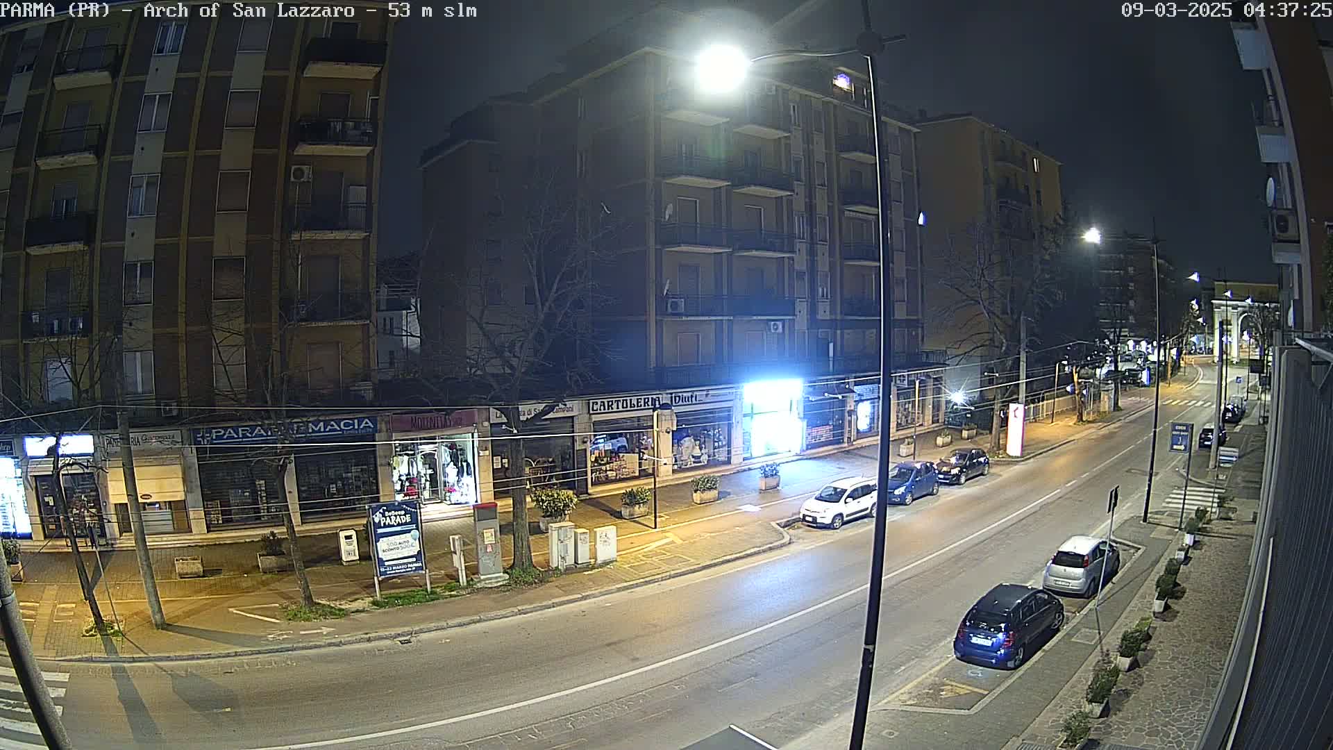 A nighttime street scene shows several parked cars along a roadway lined with shops and buildings under streetlights.