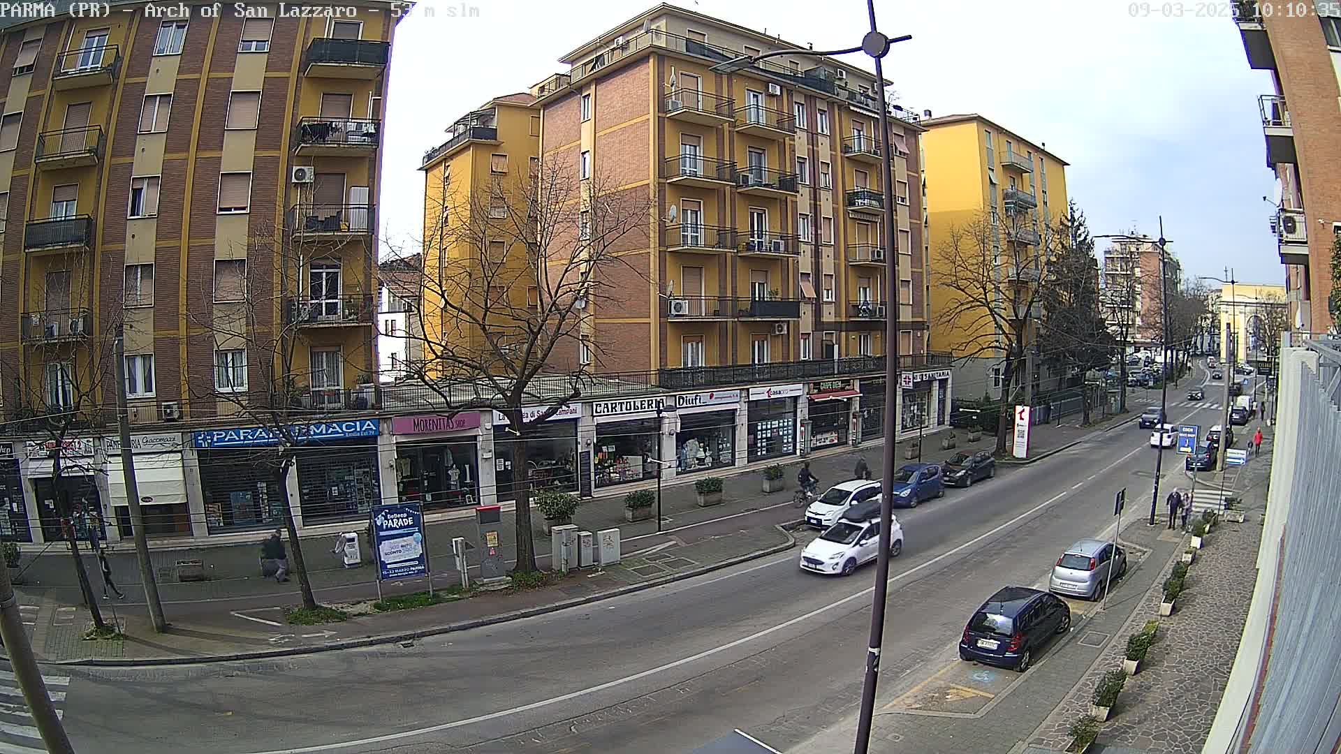 A mostly sunny street scene shows several multi-story buildings, parked cars, and pedestrians, with some bare trees lining the street.