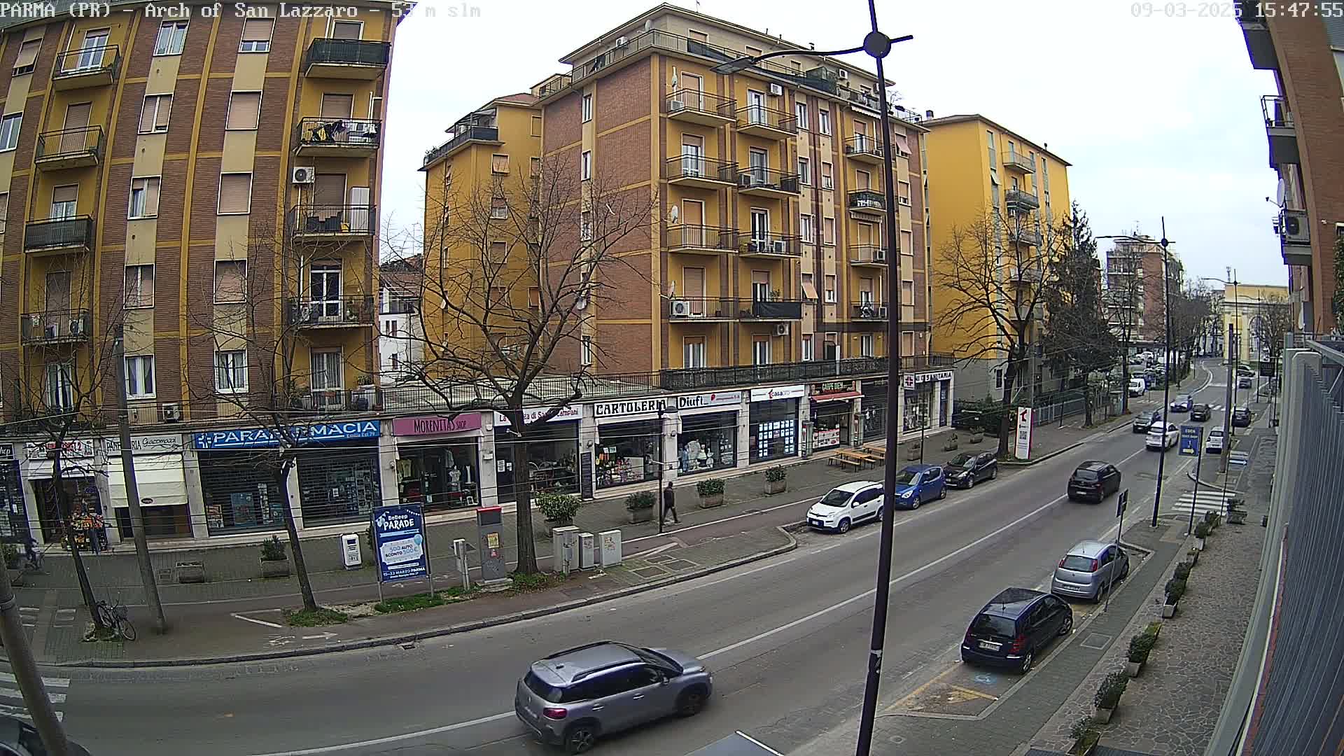 A city street scene on an overcast day shows several multi-story buildings with shops on the ground floor, cars parked along the sides of the street, and several cars driving down the street.