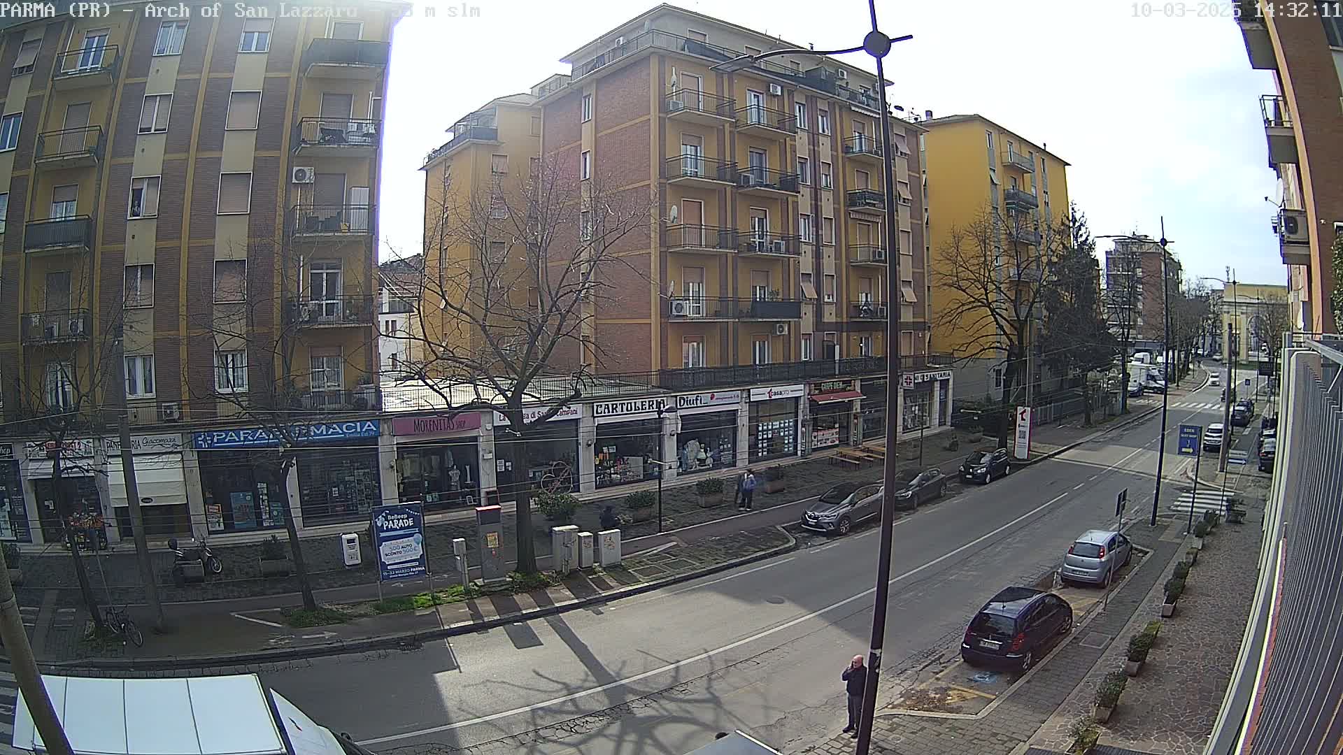 A sunny street scene shows a multi-story brick building with shops on the ground floor, several parked cars, and a few pedestrians.